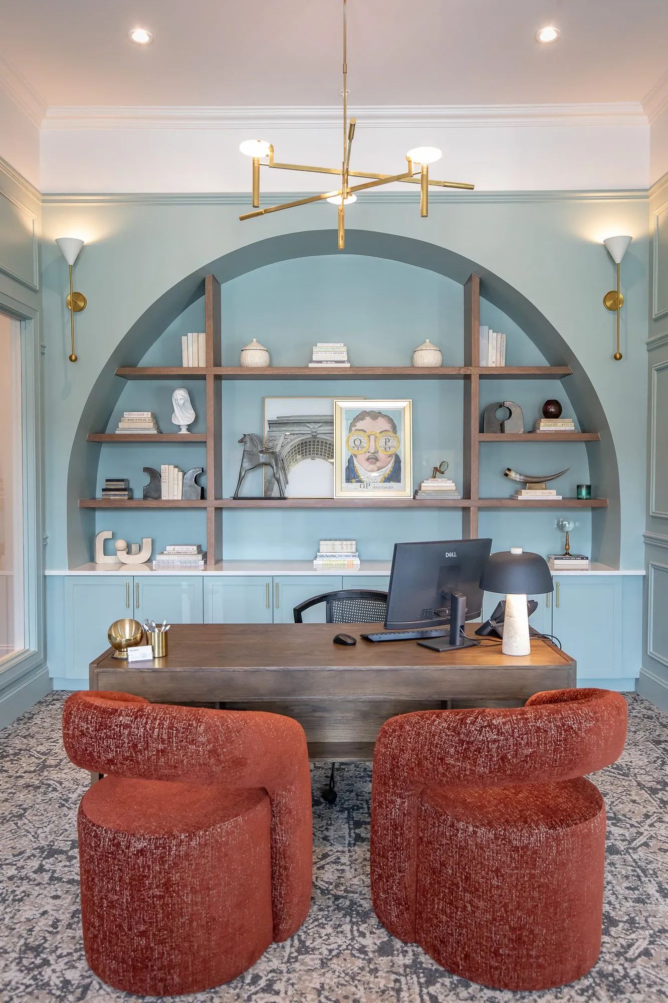 Multifamily amenity leasing office with a desk and rust boucle chairs before an arched blue niche under a brass chandelier
