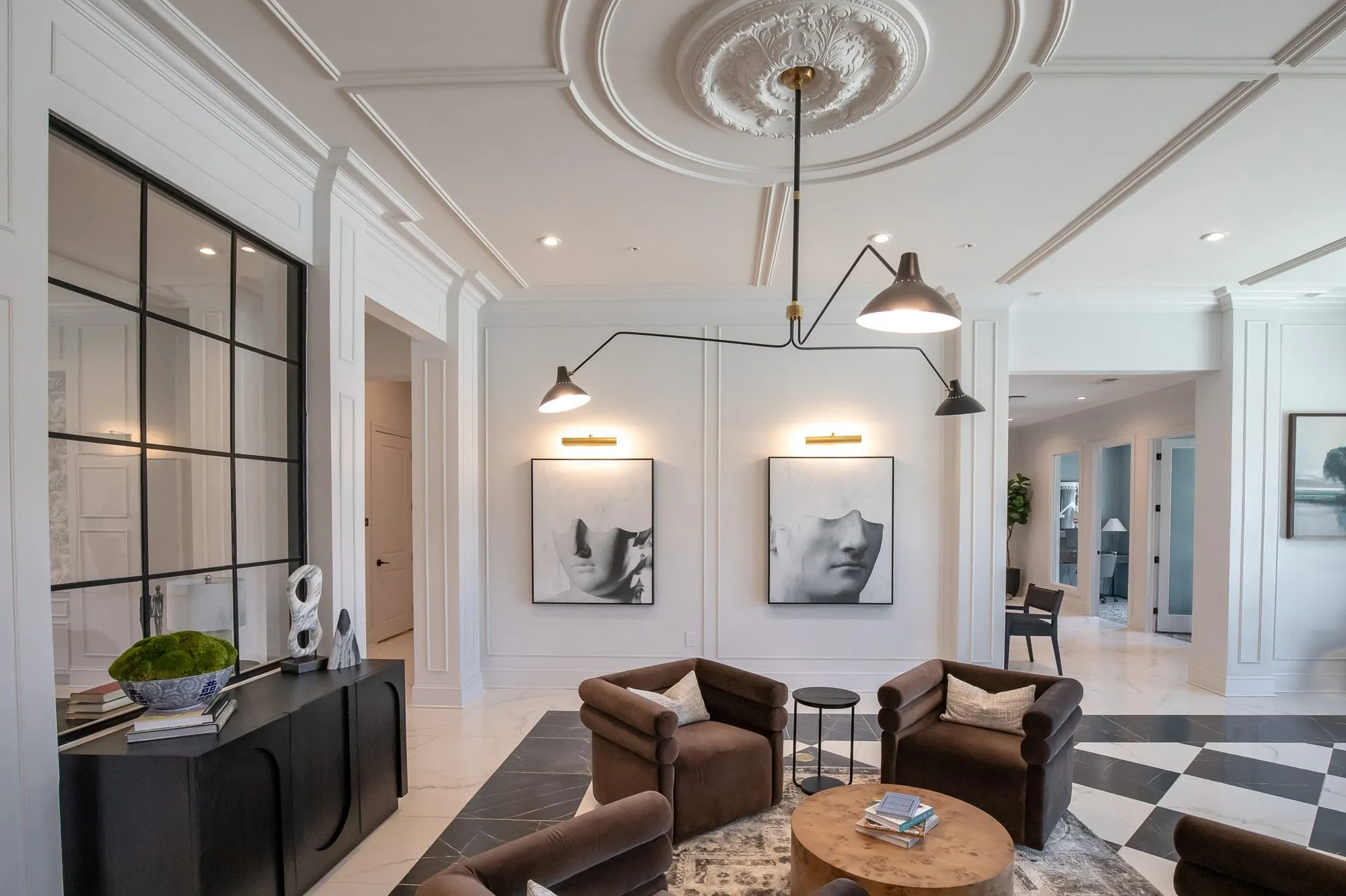 Multifamily amenity lobby lounge of brown velvet chairs under a black arm light, a fluted console and framed face prints