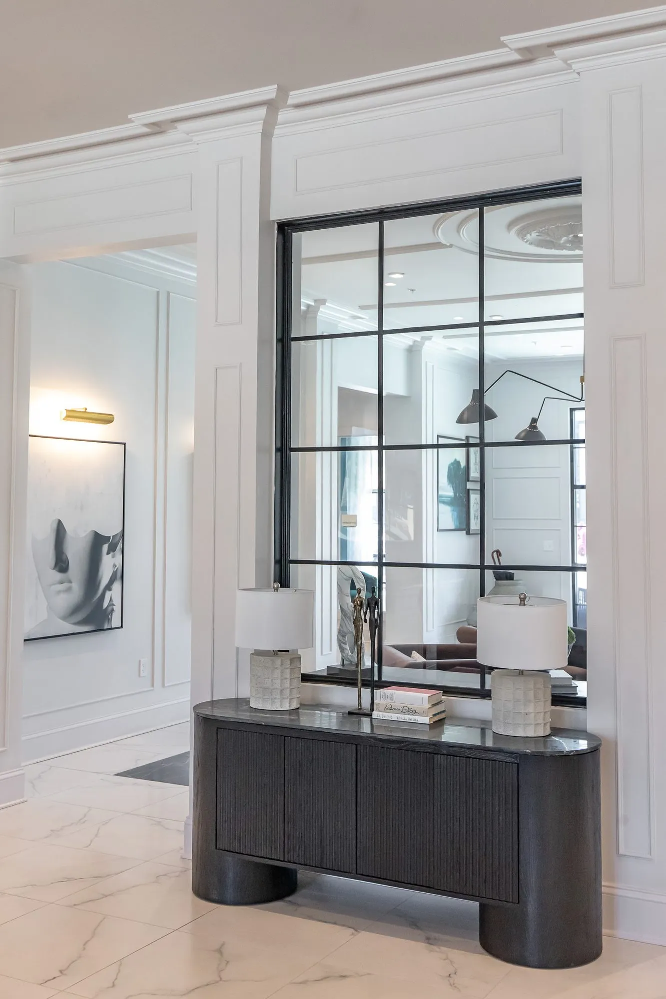 Multifamily amenity lobby lounge with a black fluted console and two lamps below a black grid mirror, a face print at left