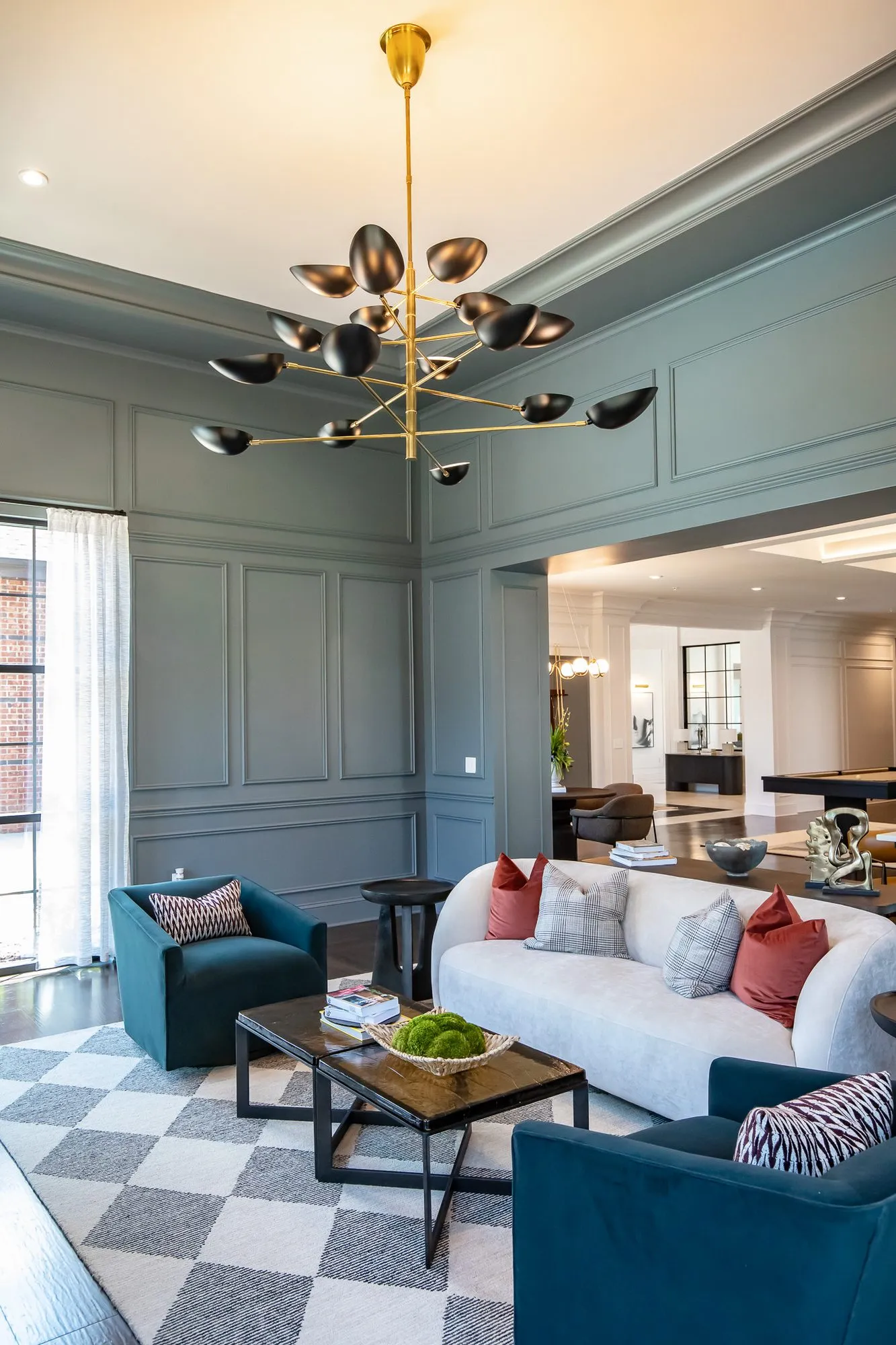 Multifamily amenity sitting room with a white curved sofa and teal chairs on a checkered rug under a brass bird chandelier