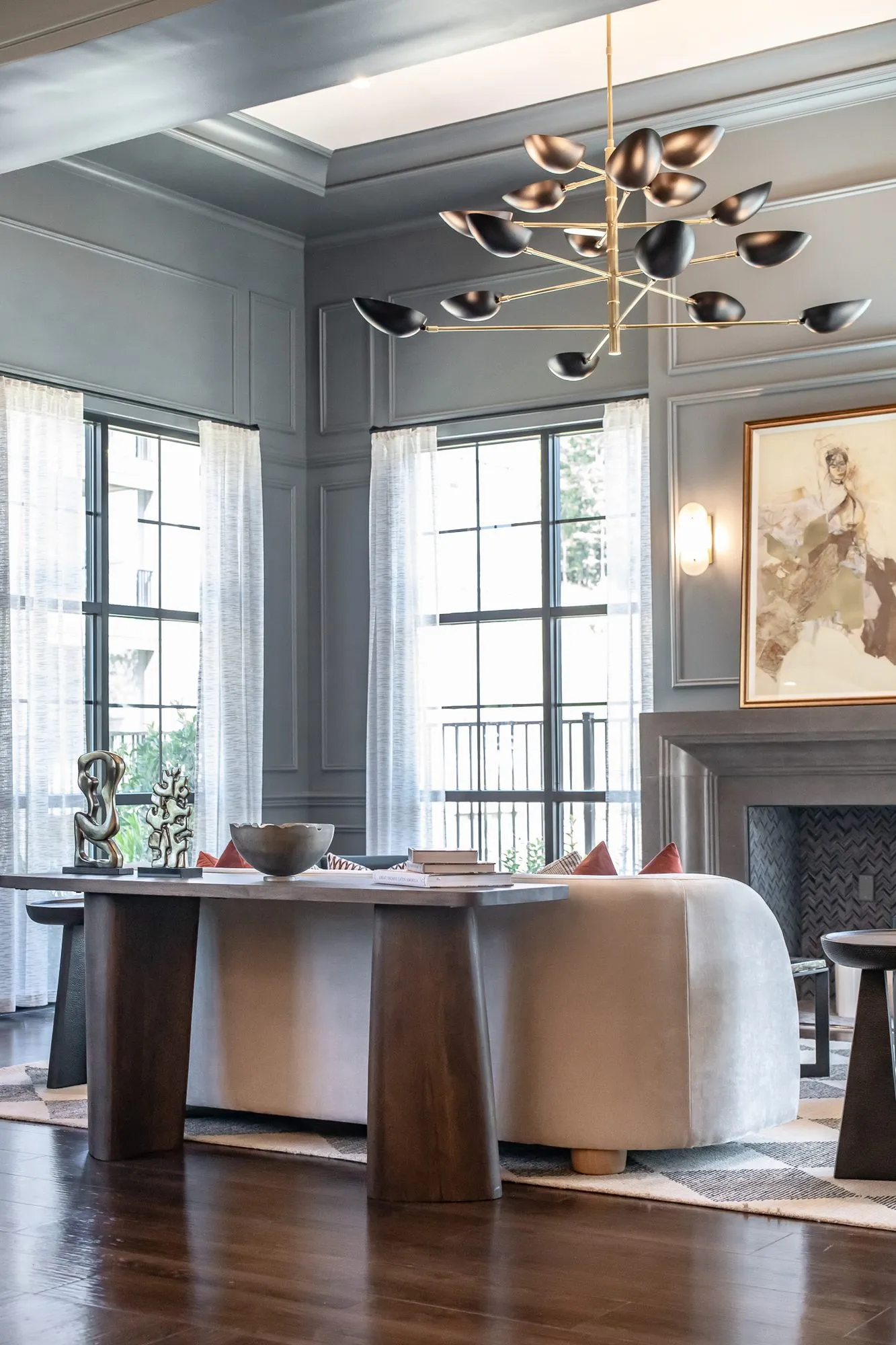 Multifamily amenity sitting room with a walnut console table of sculptures behind a curved sofa, a fireplace and tall windows