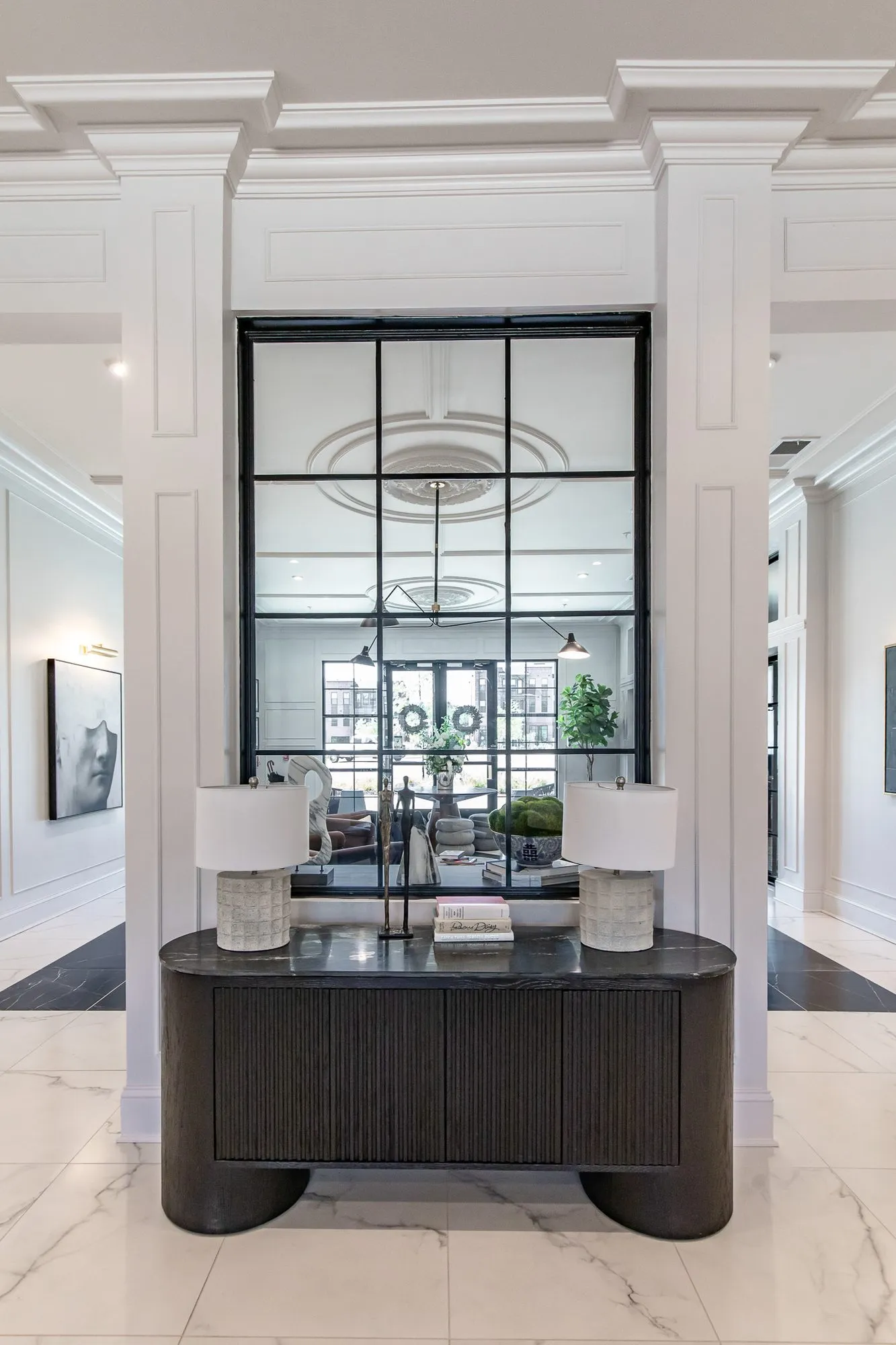 Multifamily amenity lobby lounge with a black fluted console and two lamps below a black grid mirror between white columns