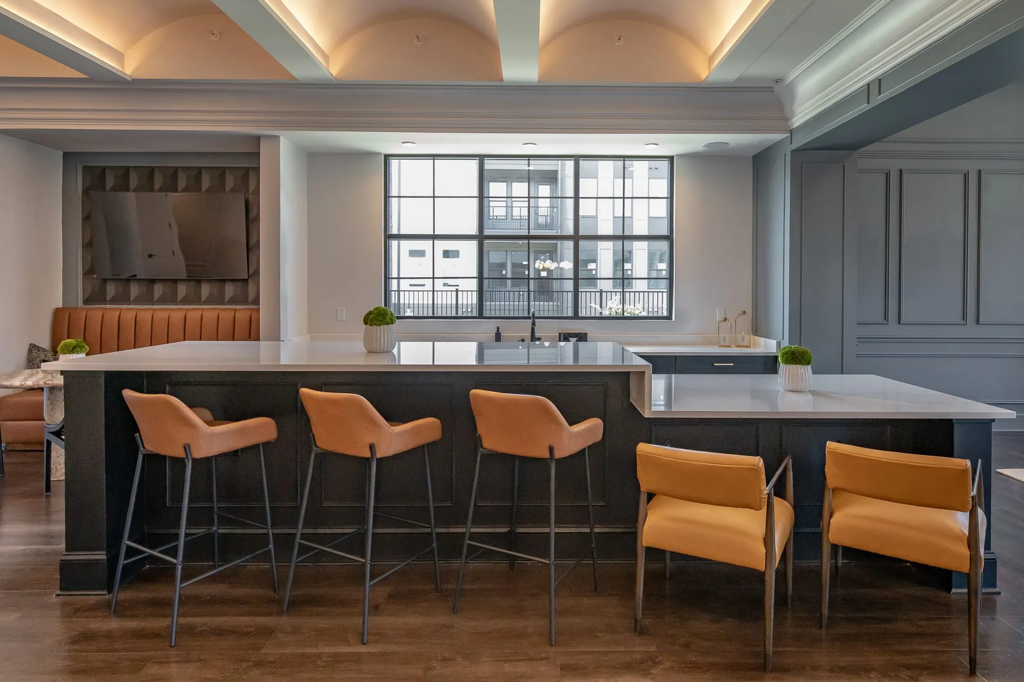 Multifamily amenity clubroom with a dark kitchen island, cognac leather stools and mustard chairs, a tufted banquette behind