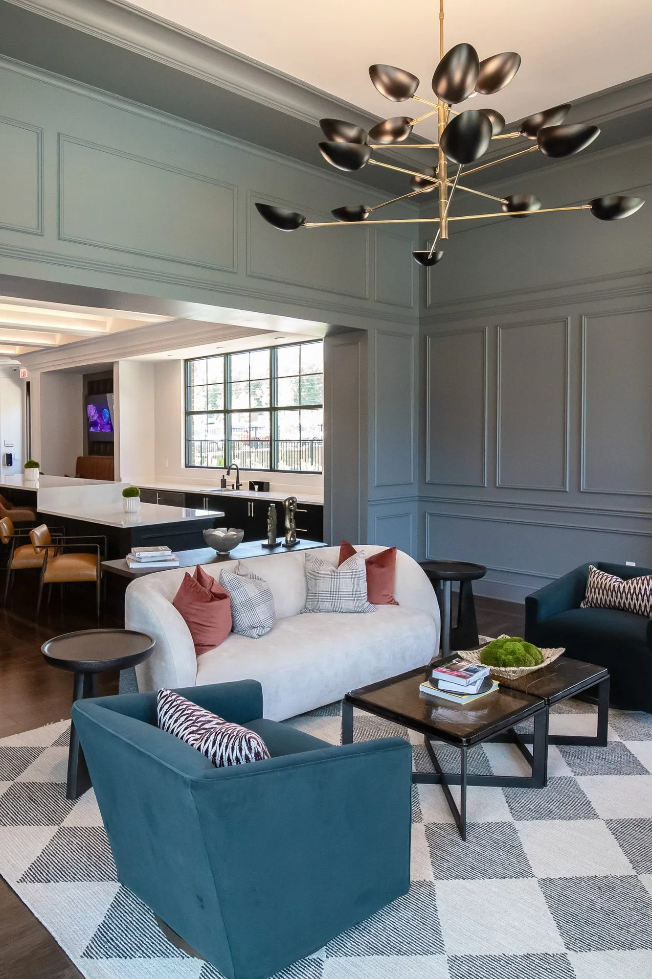 Multifamily amenity sitting room with a teal swivel chair and white curved sofa on a checkered rug, the bar beyond