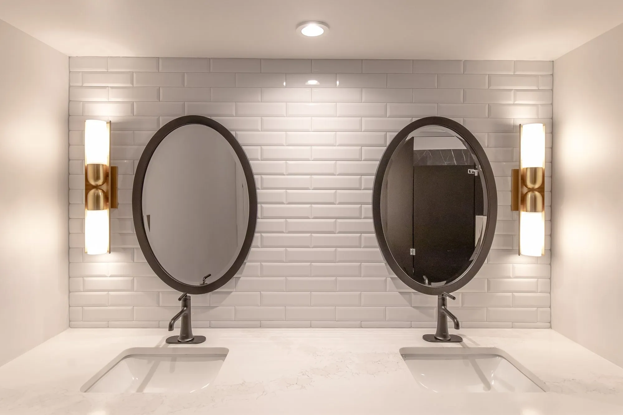 Multifamily amenity restroom with two oval black-framed mirrors and brass sconces on white beveled subway tile