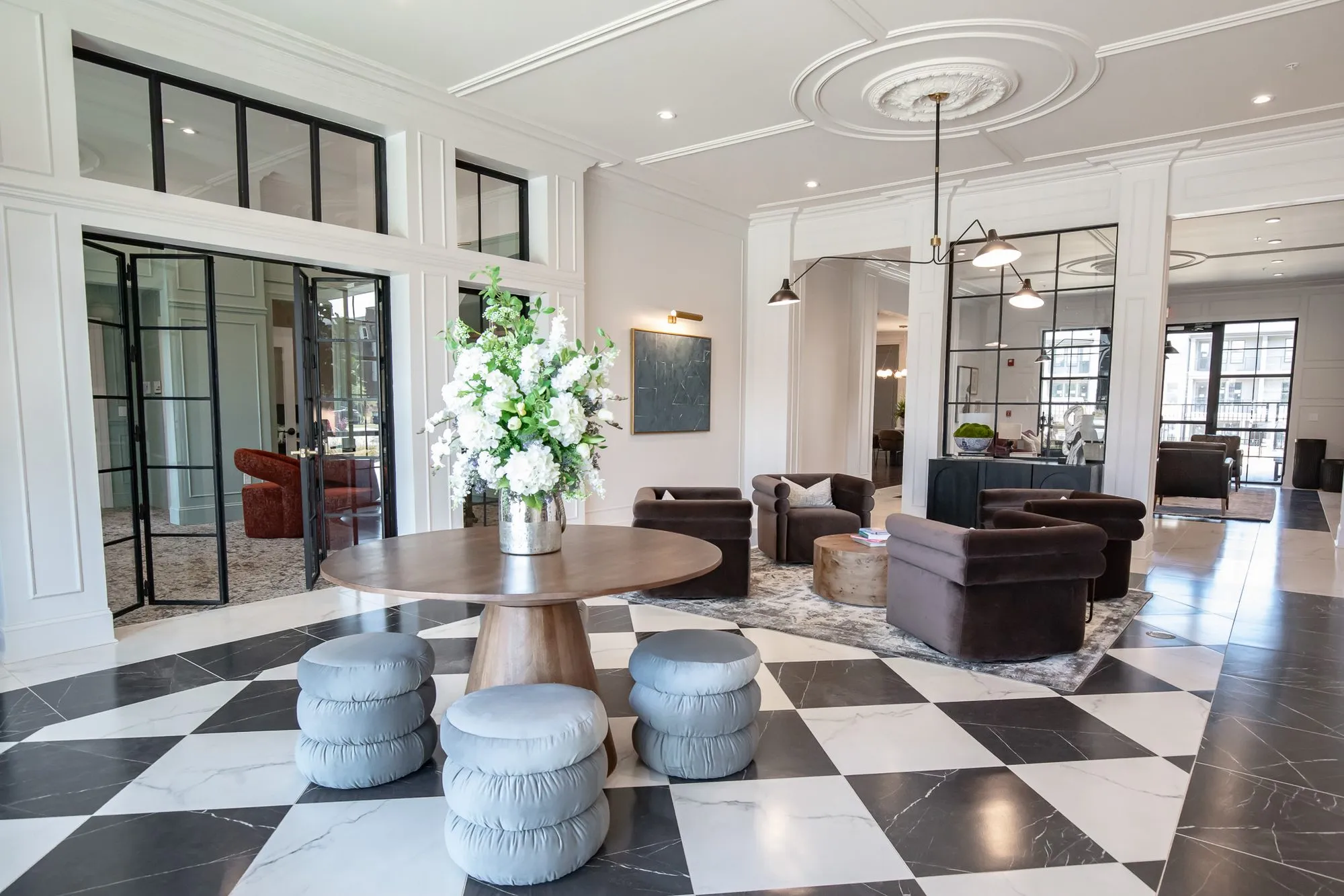 Multifamily amenity lobby toward black steel doors, a flower-topped table and puff stools on checkerboard marble