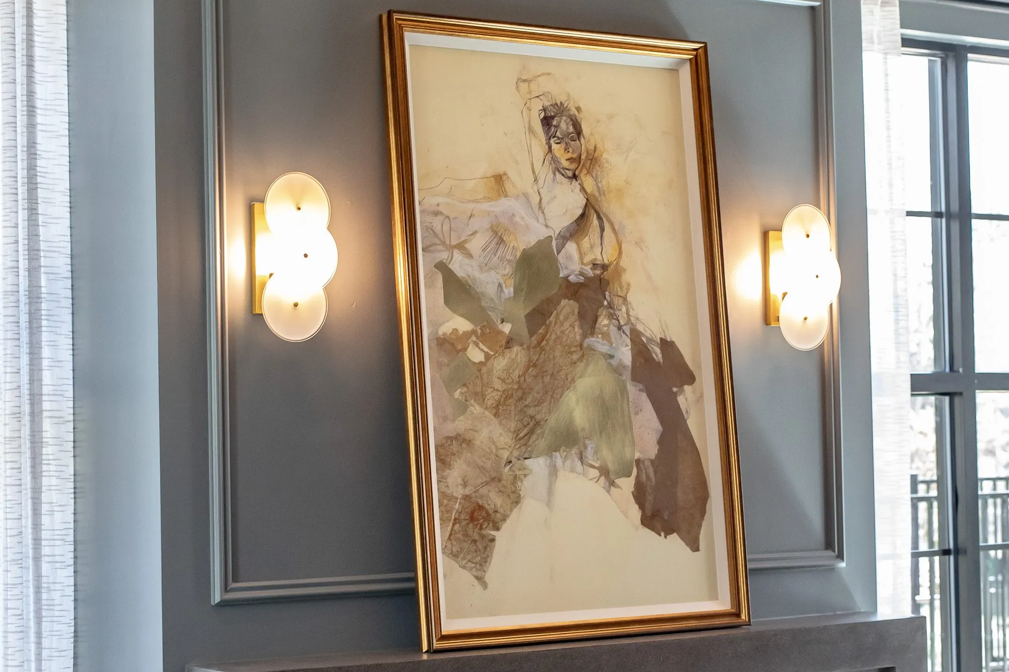 Multifamily amenity sitting room with a gilt-framed abstract figure collage between two glass disc sconces on grey panelling