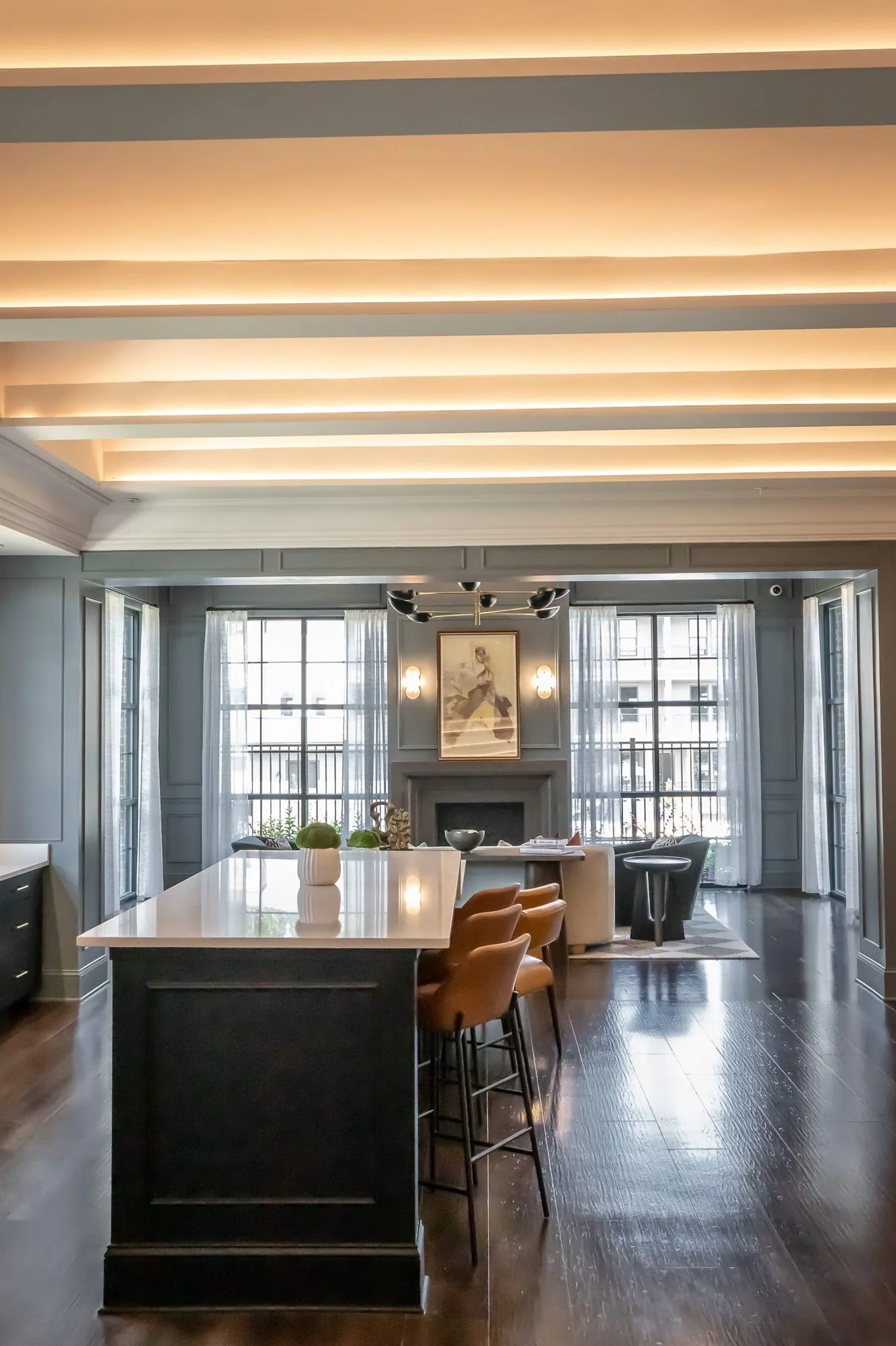 Multifamily amenity clubroom with a dark kitchen island and cognac stools under a lit tray ceiling, the fireplace room beyond
