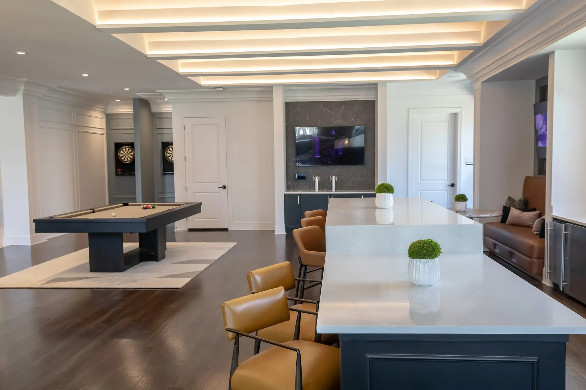 Multifamily amenity clubroom with a long white island and cognac leather stools, the billiards table and dartboards beyond