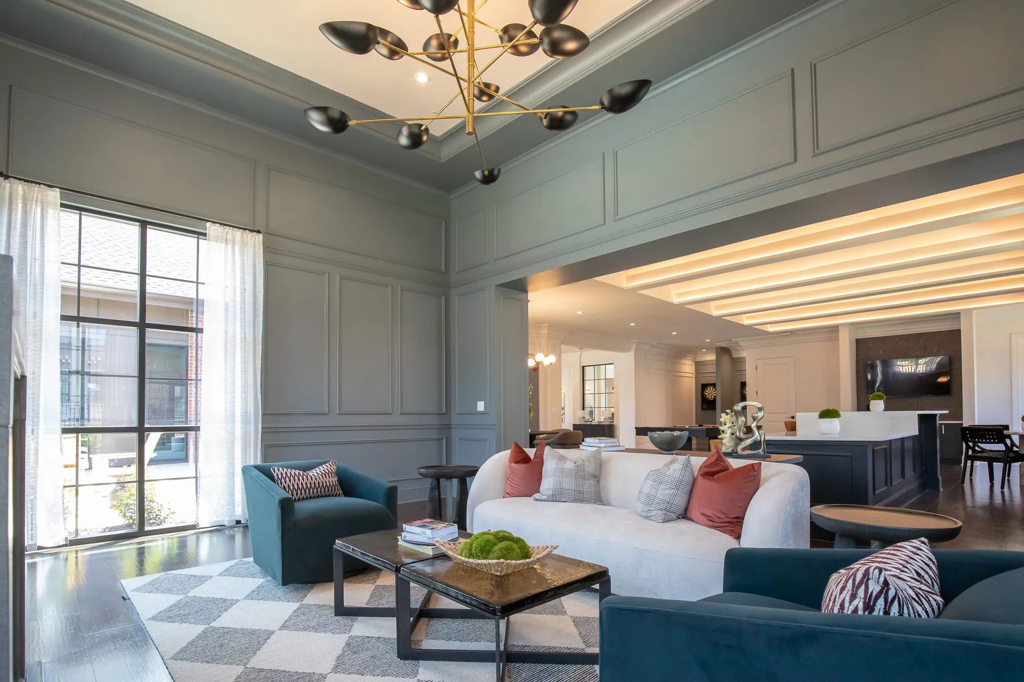 Multifamily amenity grey panelled lounge with a white curved sofa and teal chairs under a brass bird chandelier, bar beyond