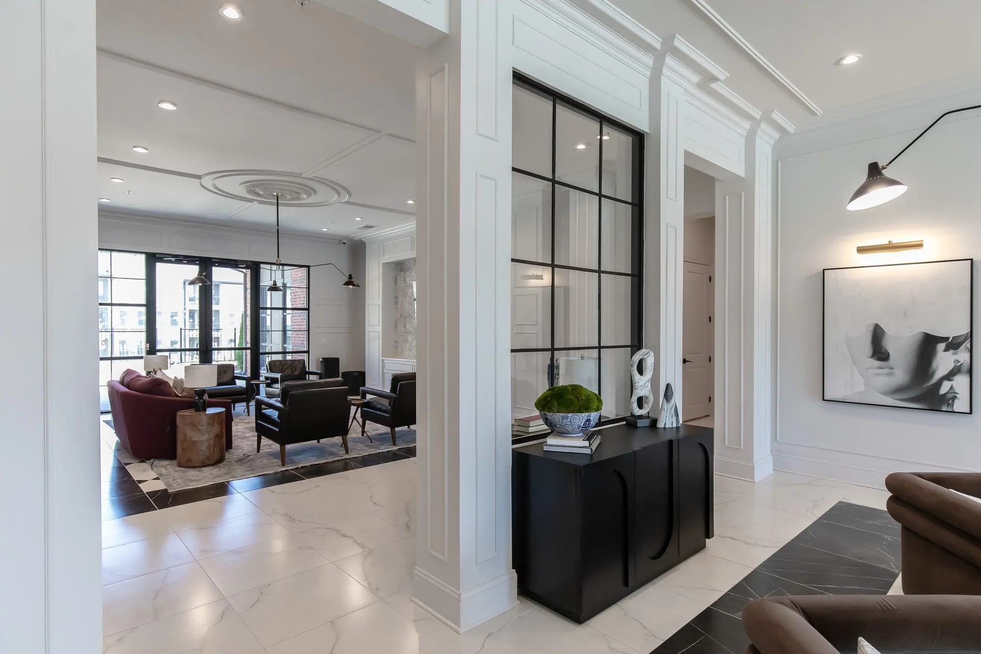 Multifamily amenity lobby corridor with a black console and moss bowl by black steel glazing, a burgundy sofa beyond