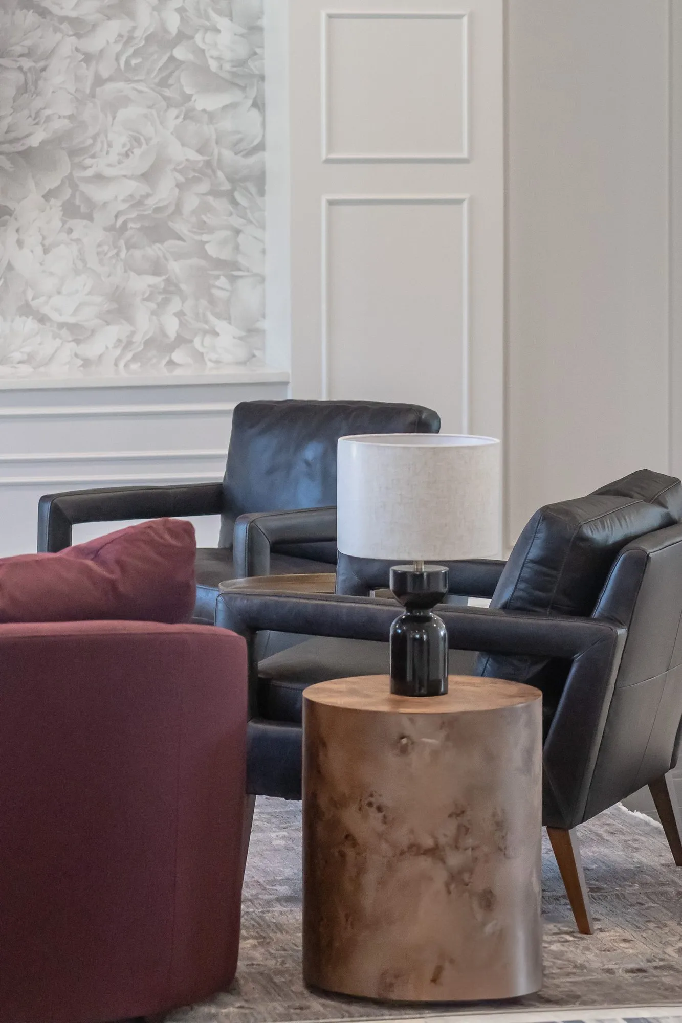 Multifamily amenity clubroom with black leather armchairs and a burl side table by a burgundy sofa, floral relief wallpaper