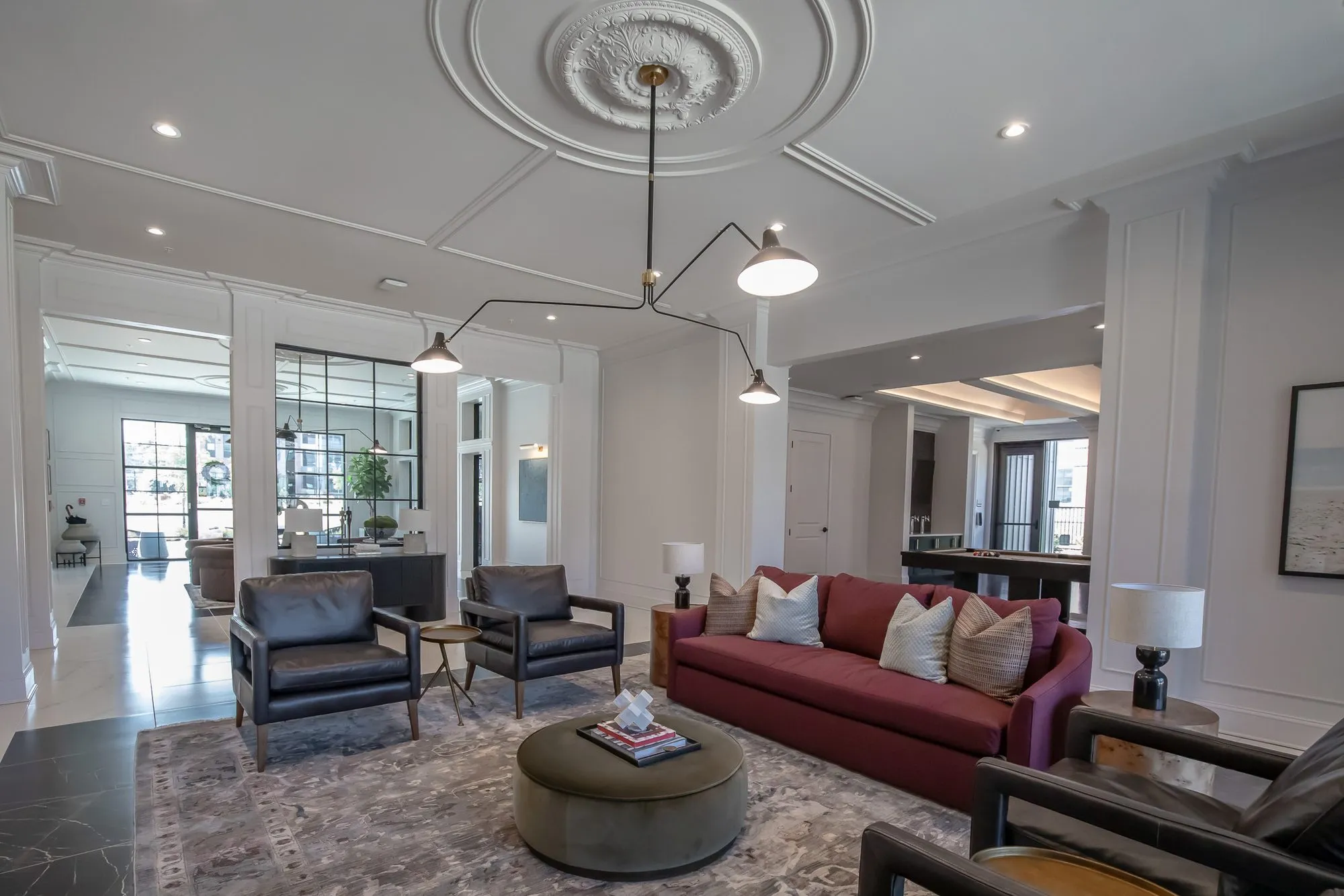Multifamily amenity clubroom with a burgundy sofa and black leather chairs around an olive ottoman under a black arm light