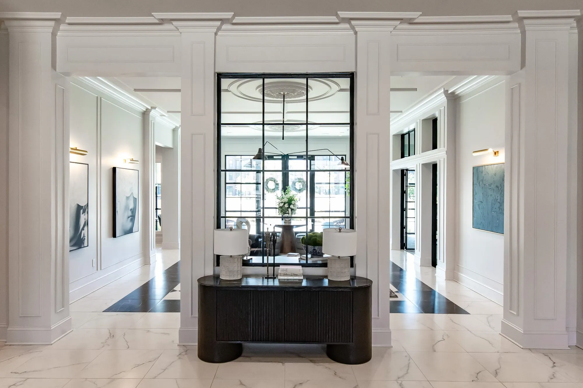 Multifamily amenity lobby lounge with a black fluted console and lamps below a black grid mirror, framed by white columns