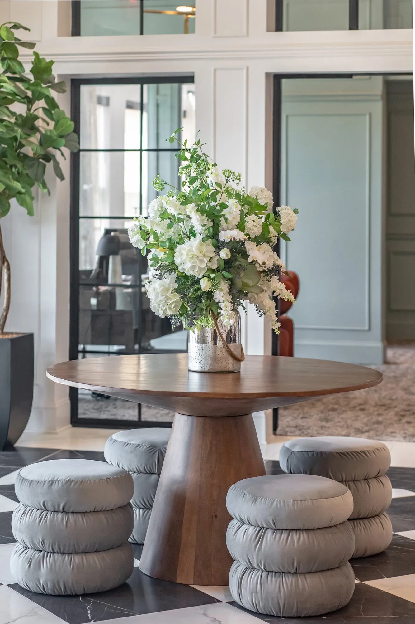 Multifamily amenity lobby with a round wood table of white flowers ringed by grey puff stools, black steel doors behind