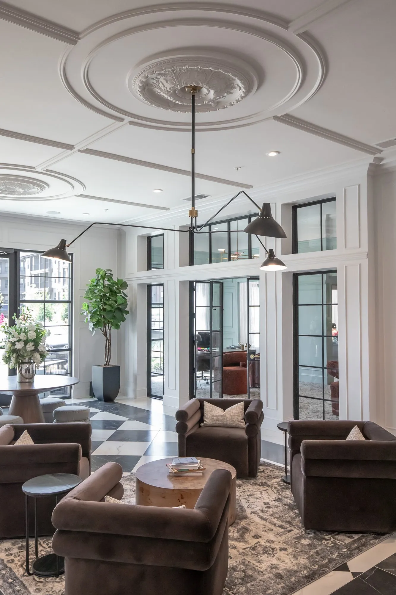 Multifamily amenity lobby lounge with brown velvet chairs under a black arm light and medallion, tall black framed windows