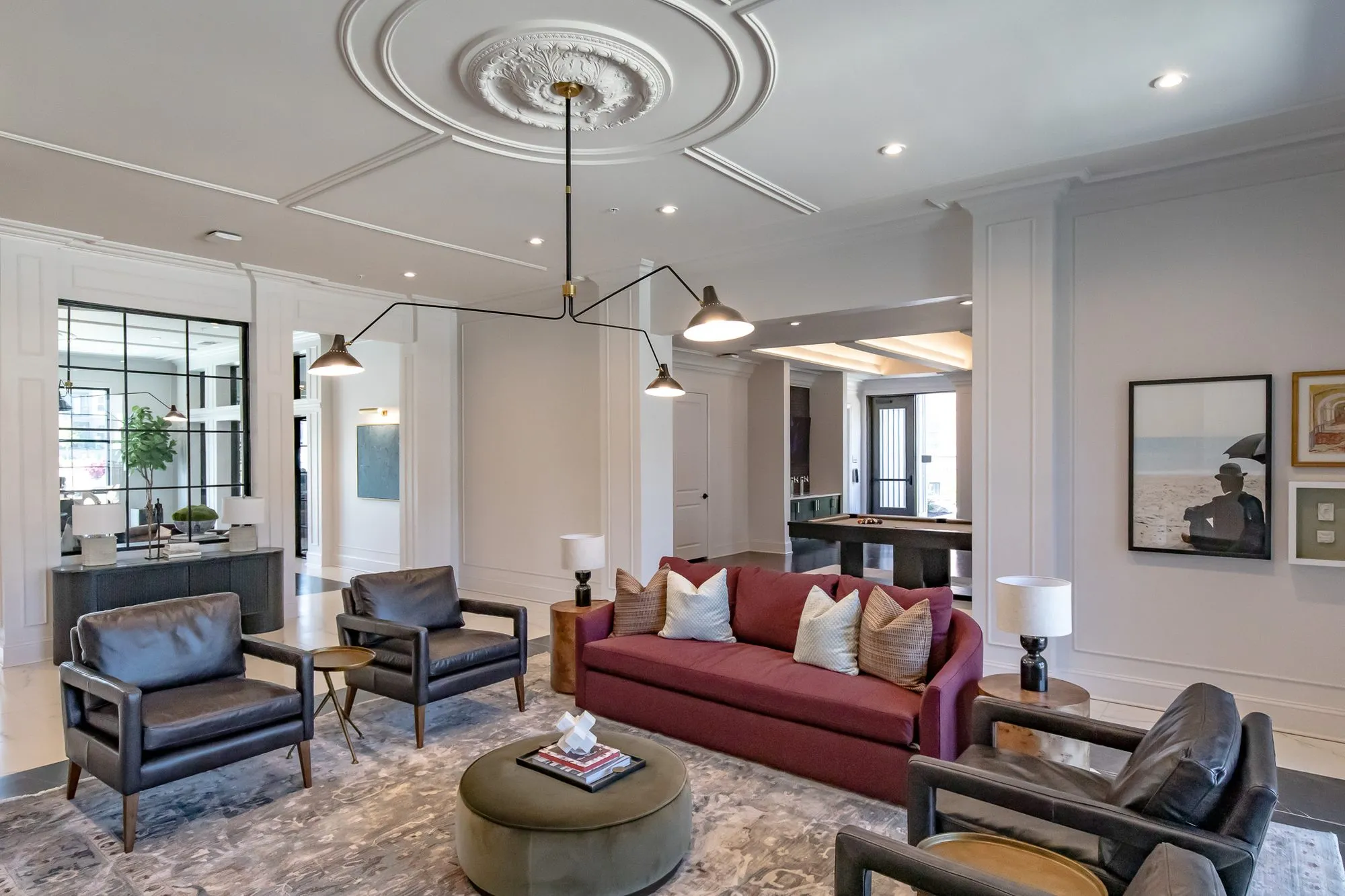 Multifamily amenity clubroom with black leather armchairs and a burgundy sofa around an olive ottoman, billiards room beyond
