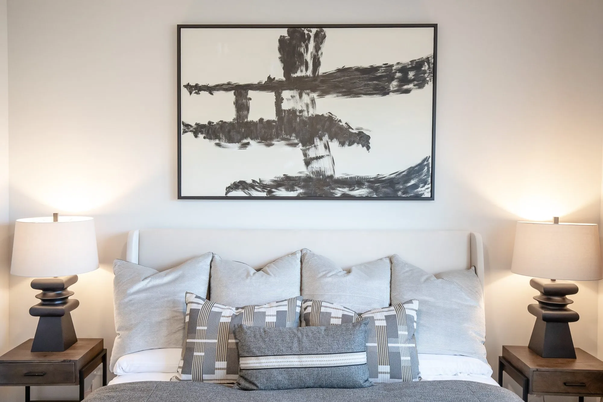 Multifamily apartment bedroom with grey and striped pillows between black stacked lamps below a black brushstroke abstract