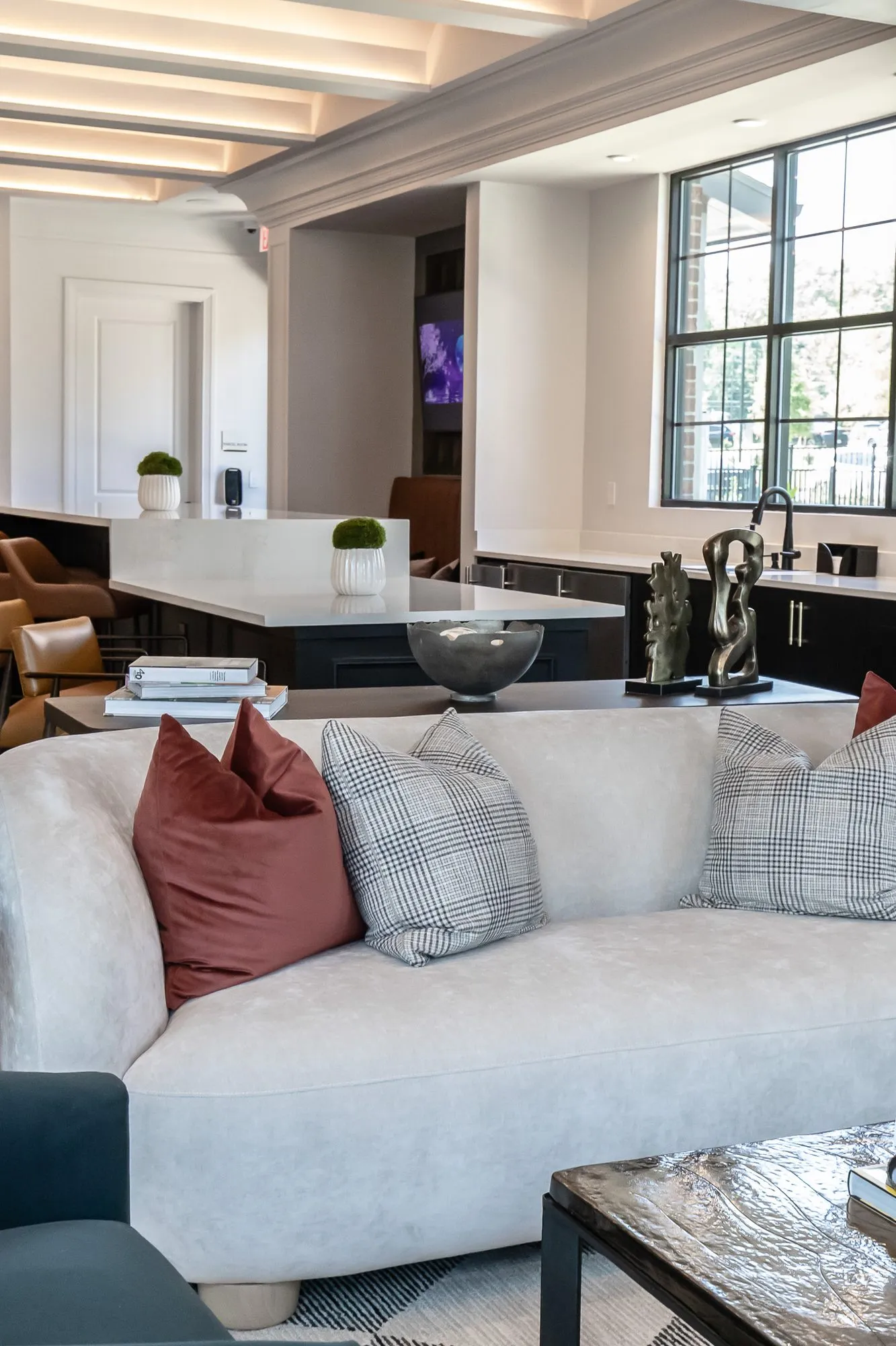 Multifamily amenity sitting room seen over a white curved sofa with rust and plaid pillows to the kitchen island and stools