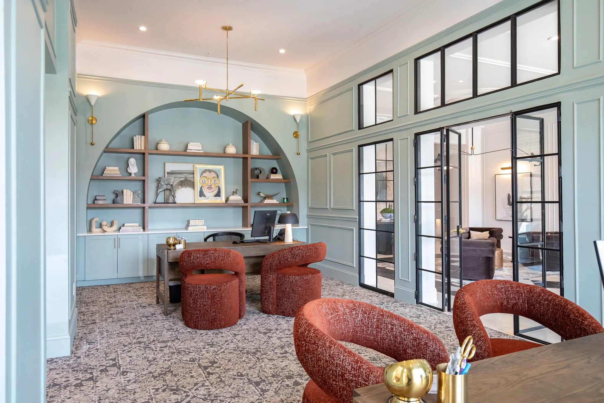 Multifamily amenity study with arched sage built-in shelving, a desk and rust boucle chairs, steel framed glass doors