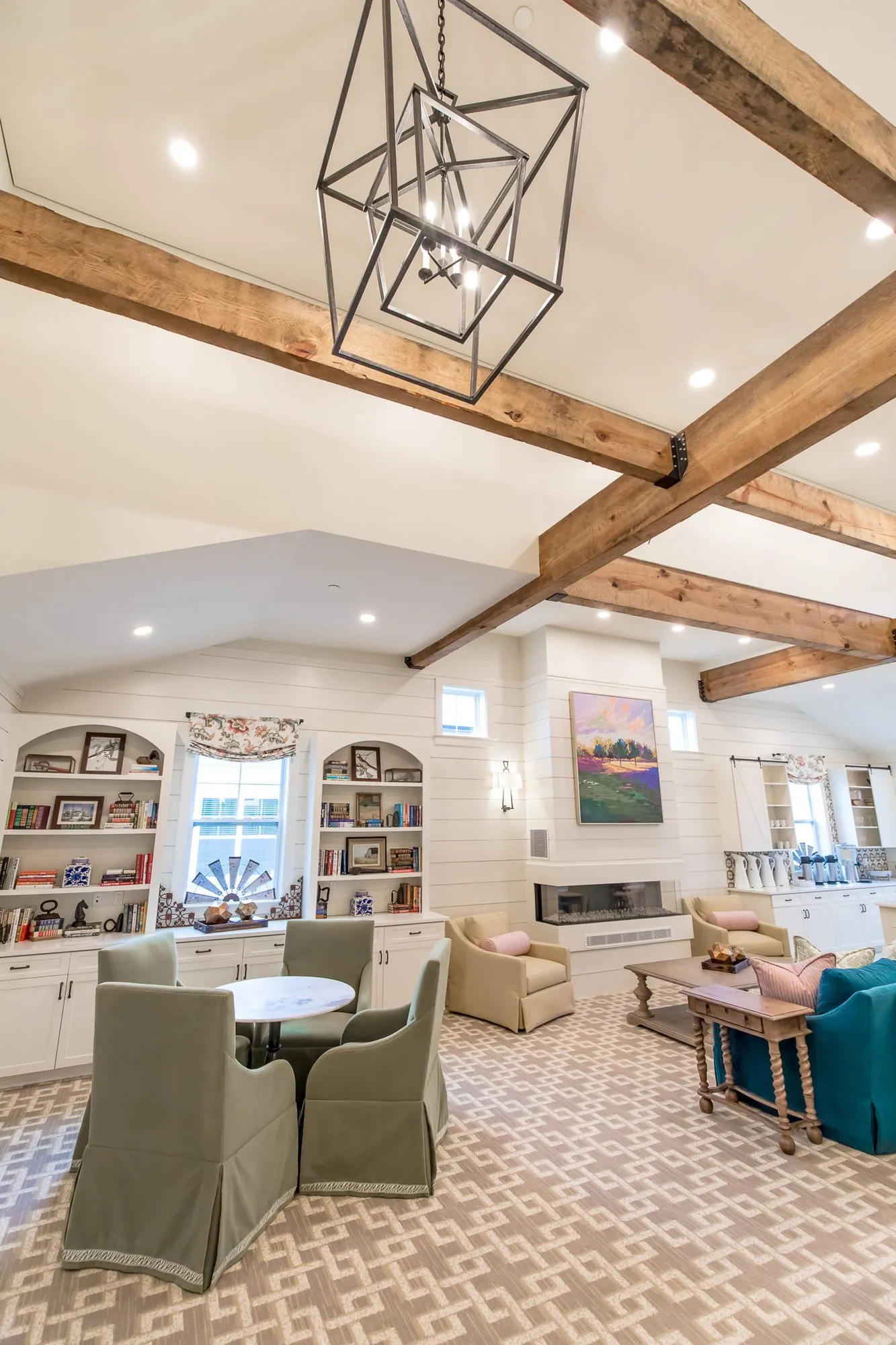 Senior living lounge with sage skirted chairs at a round table under timber beams and a black cage pendant, arched bookcases