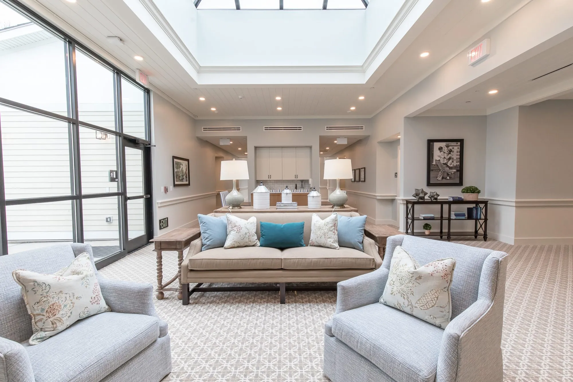 Senior living sitting area under a skylight, pale blue armchairs and a linen sofa beside a black framed window wall