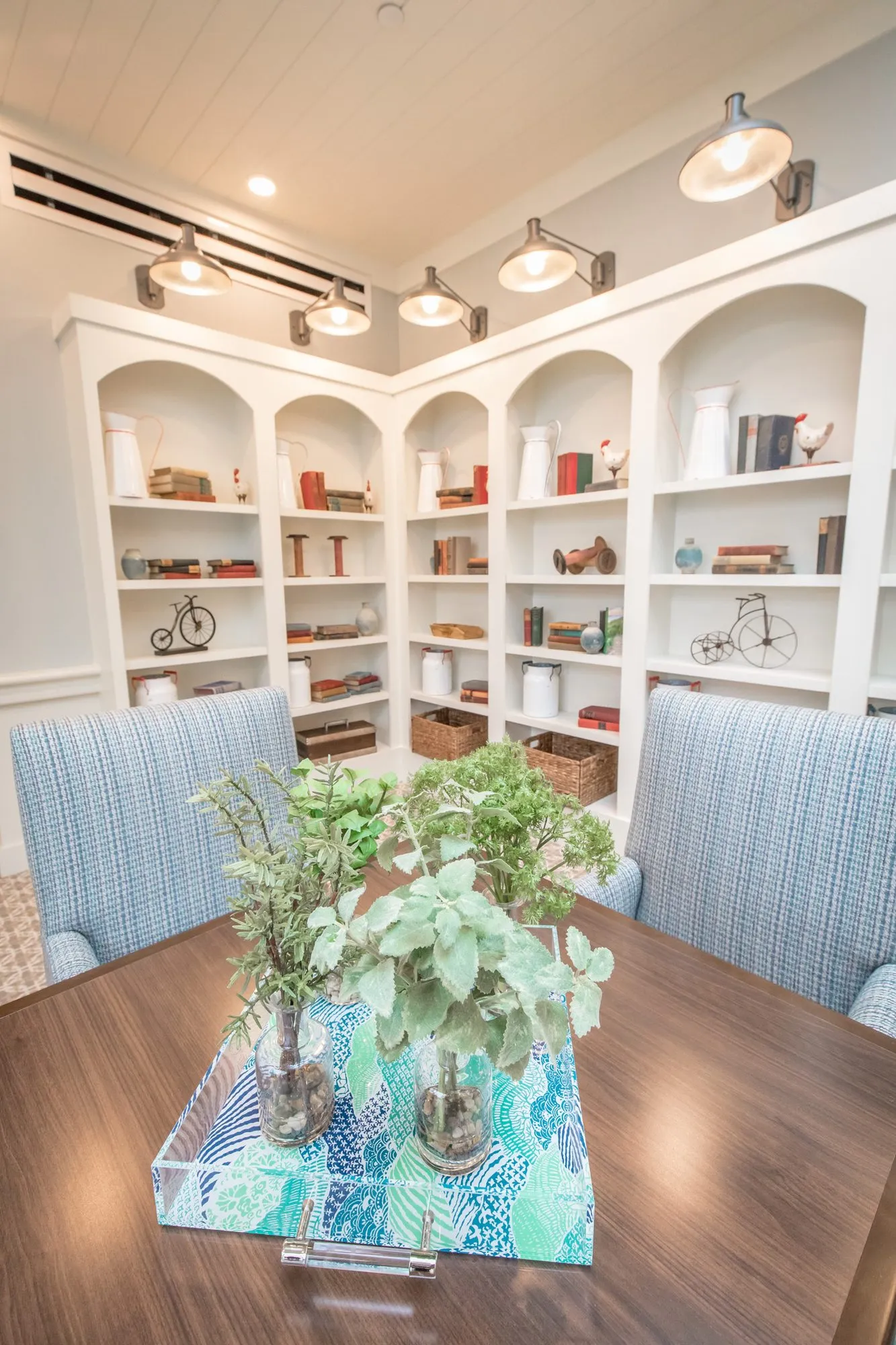 Senior living interior detail: wood table with glass jars of greenery on a blue tray before arched bookcases and barn sconces