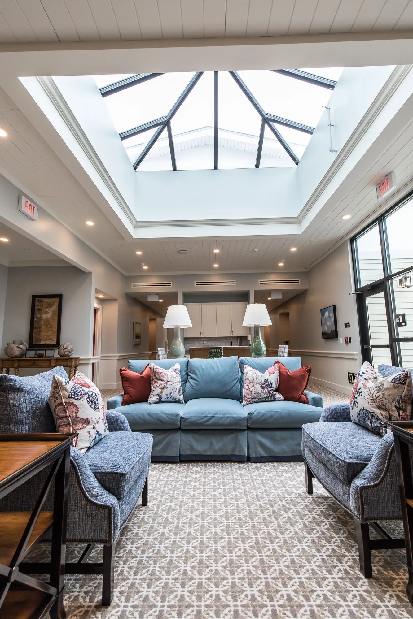 Senior living atrium lounge with a blue skirted sofa and red pillows between blue armchairs below the pyramid skylight