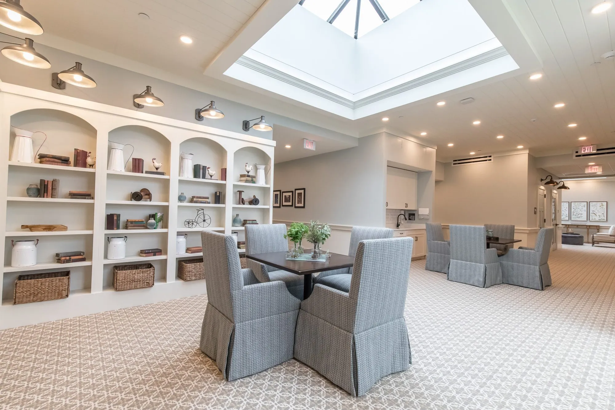 Senior living library with arched white bookcases and barn sconces, grey skirted chairs at square tables under a skylight