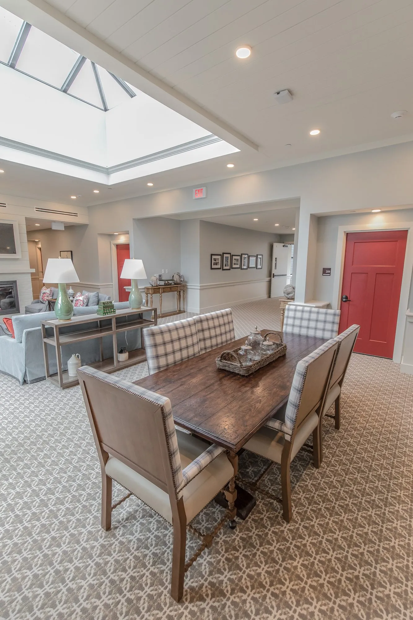 Senior living dining area with plaid-backed chairs at a table under a skylight, a red door and a lamp-lit console beyond