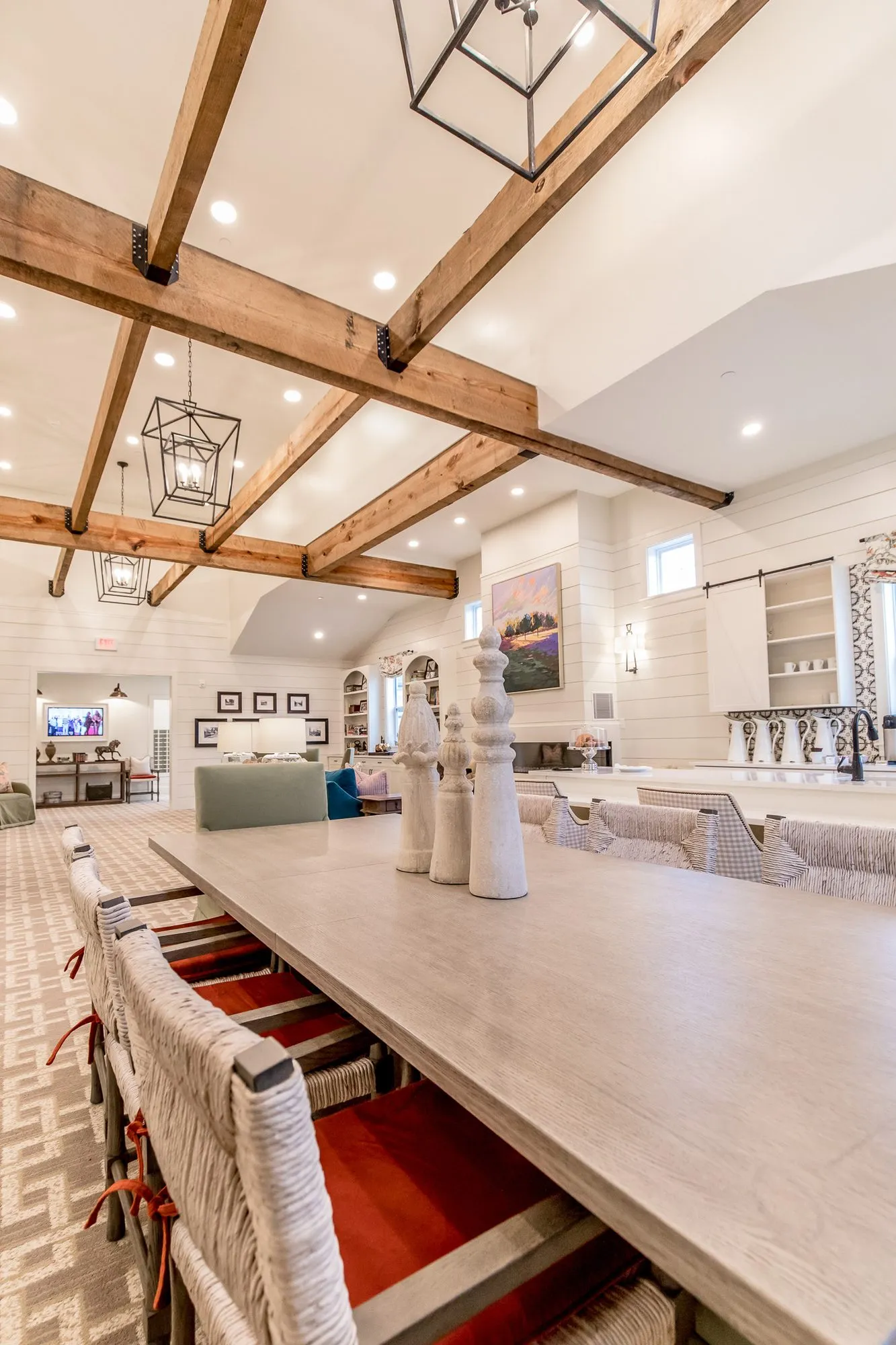 Senior living interior detail: long whitewashed farm table with woven-back chairs under timber beams and cage pendants