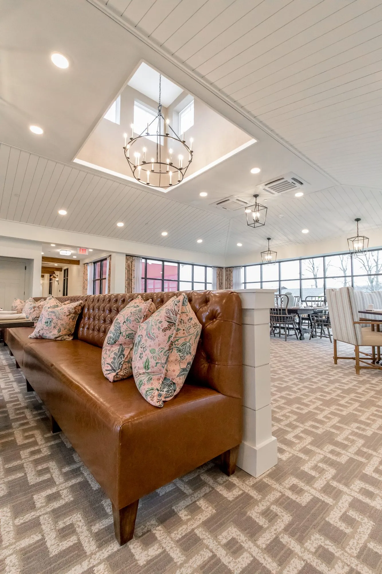 Senior living dining room with a tufted cognac leather banquette and floral pillows under a skylight and ring chandelier