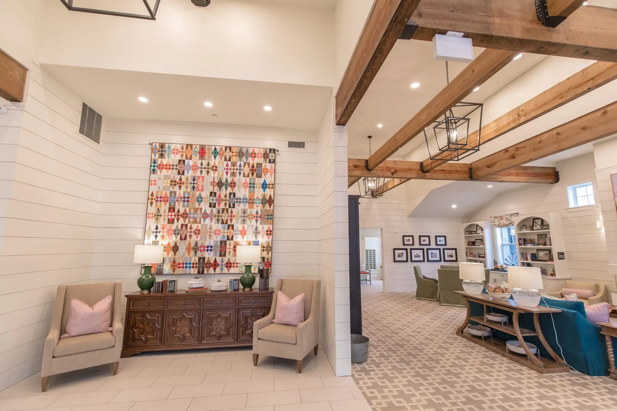 Senior living entry with a patchwork quilt above a carved sideboard between two linen armchairs, the great room beyond