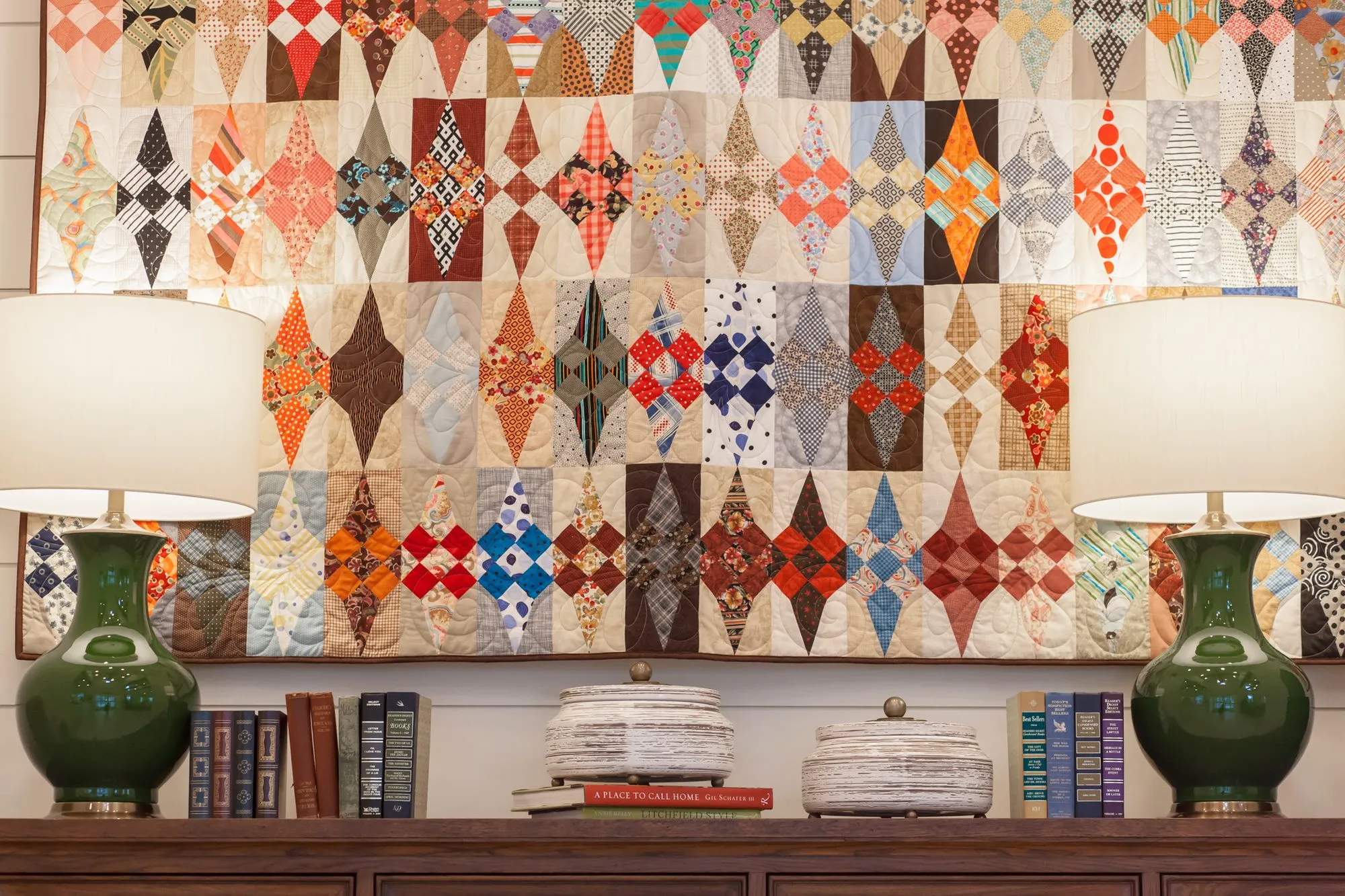 Senior living interior detail: diamond-pattern patchwork quilt above green ceramic lamps and stacked books on a sideboard