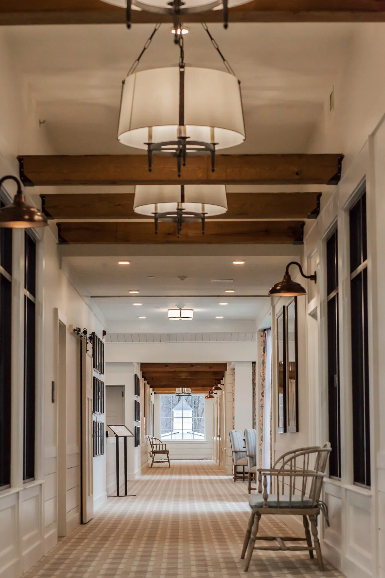 Senior living corridor under timber cross beams and white drum pendants, a spindle-back chair beside black framed doorways