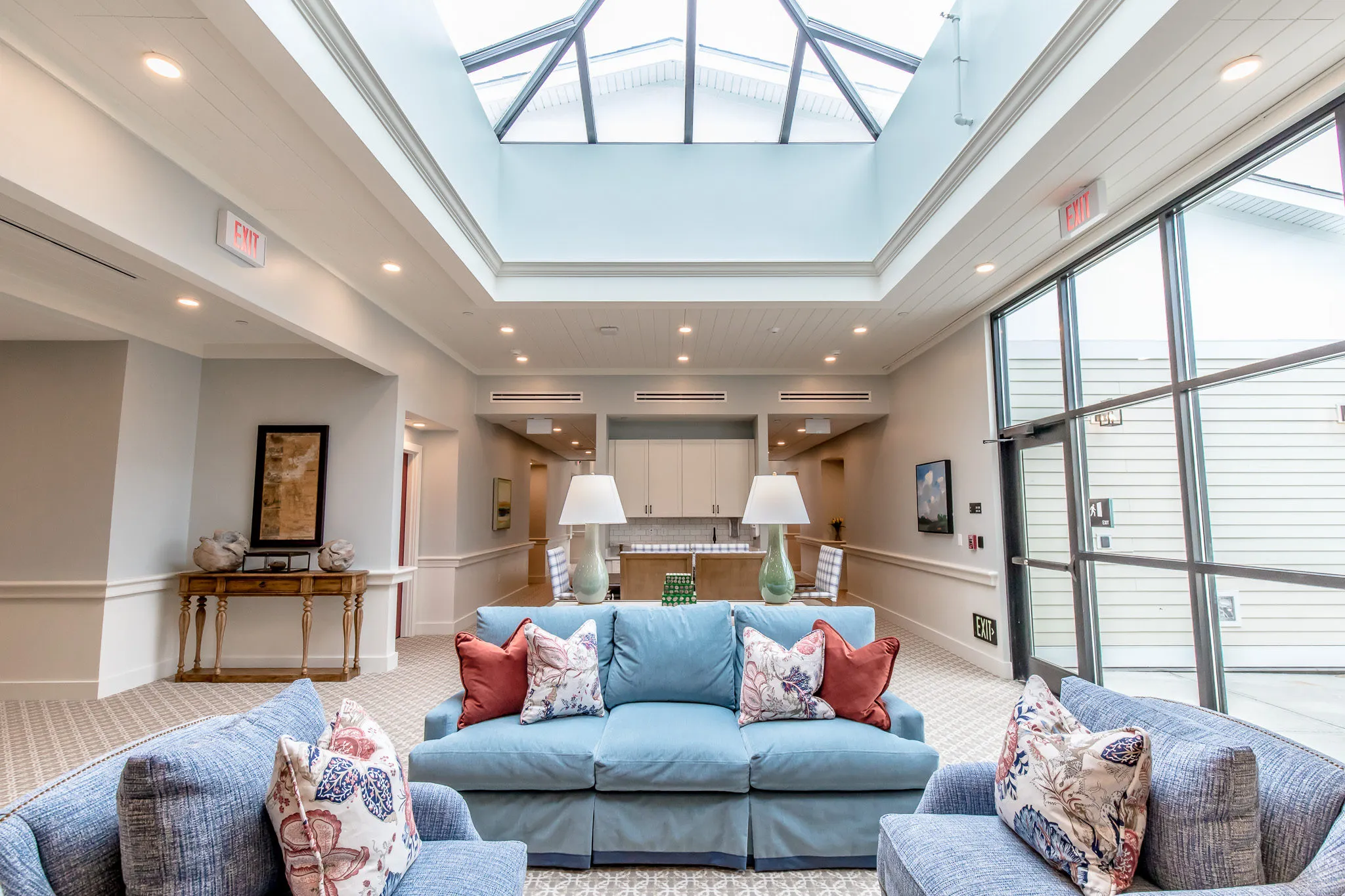 Senior living atrium lounge with blue sofas and floral pillows under the pyramid skylight beside the window wall