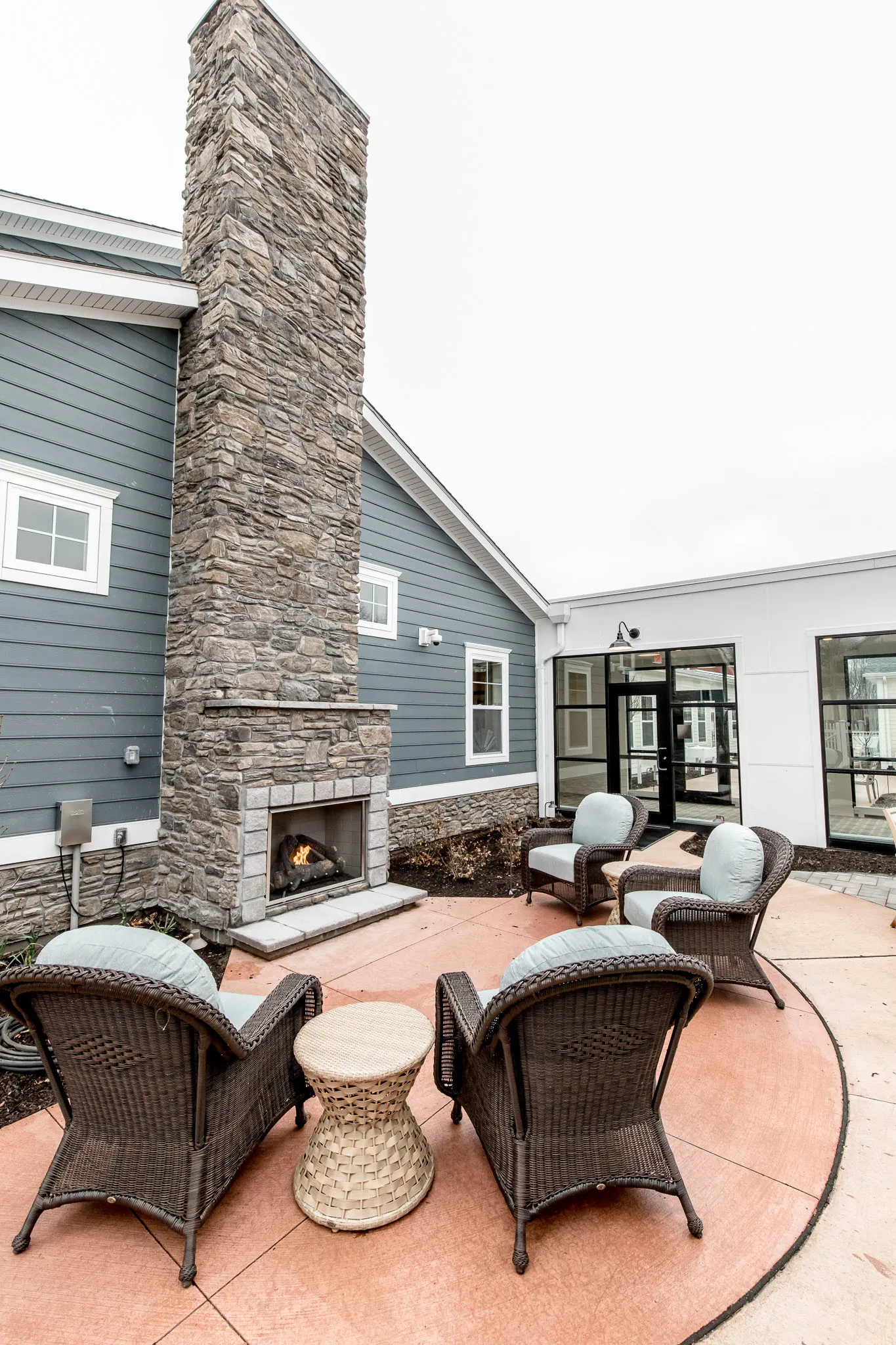 Senior living courtyard with a stacked stone chimney and outdoor fireplace, woven armchairs on a terracotta-toned patio