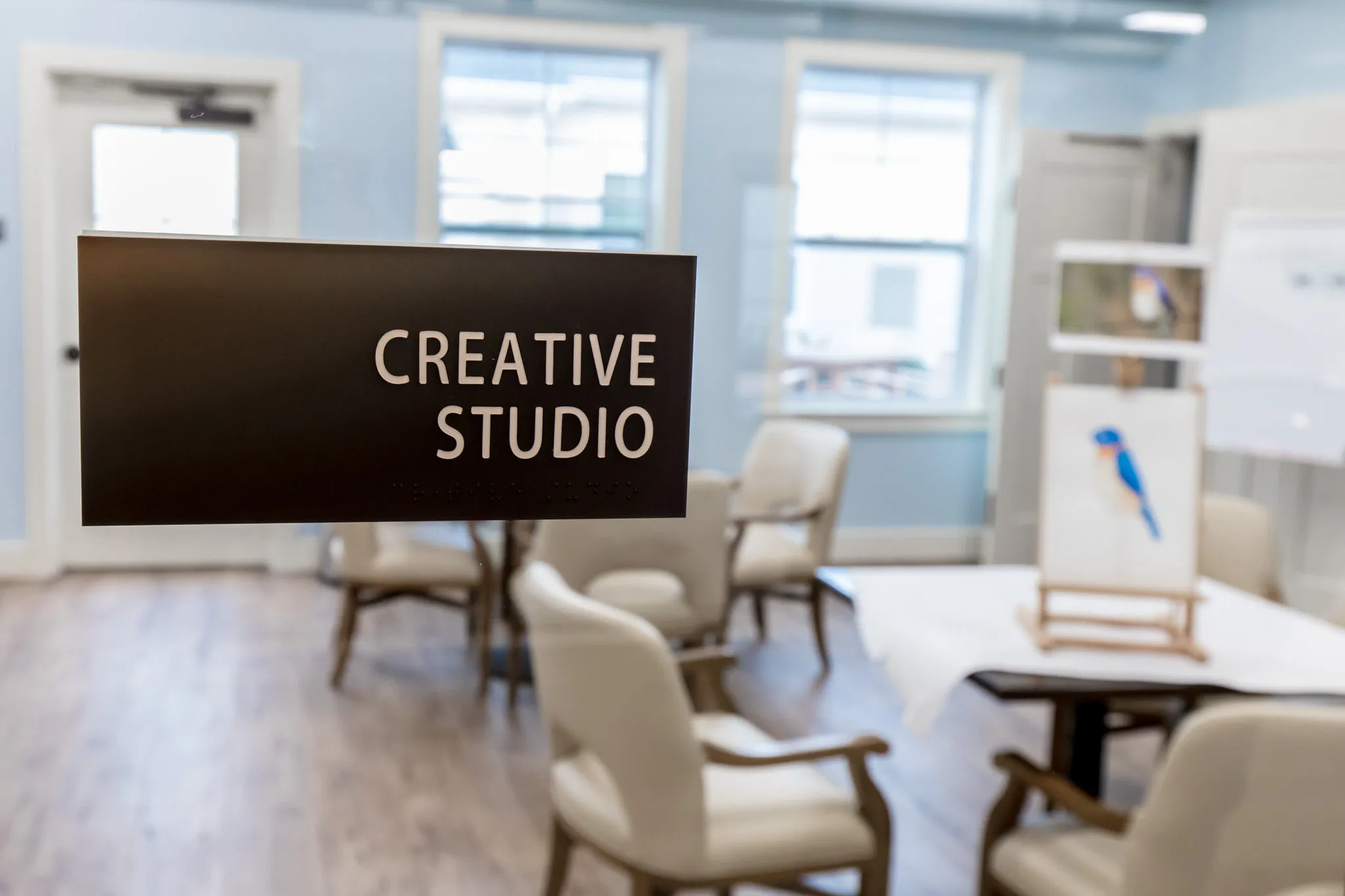 Senior living creative studio with an easel and worktables in the daylit corner room below the studio sign