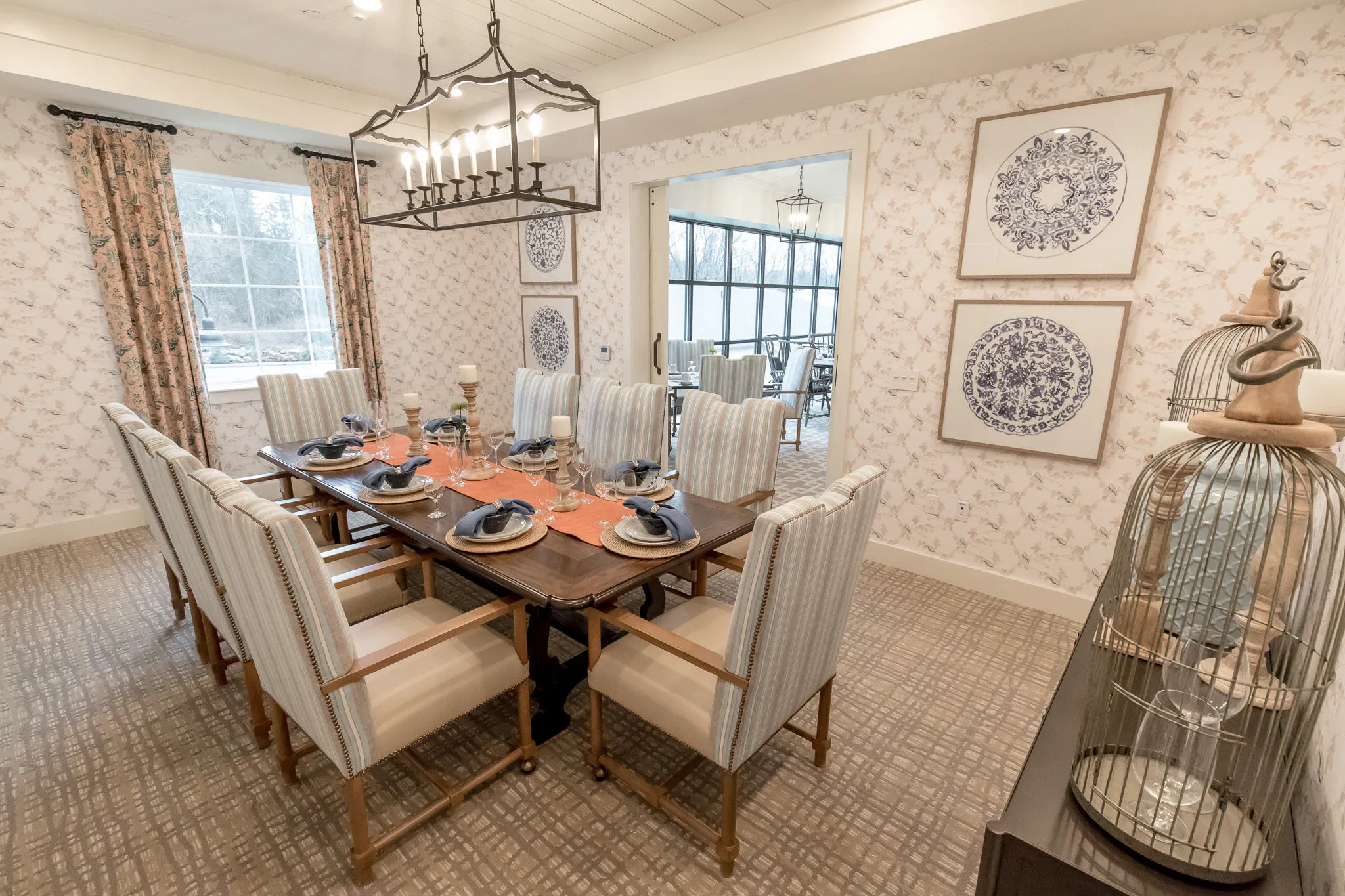 Senior living private dining room with linen chairs around a set table under a lantern chandelier beside a vintage birdcage
