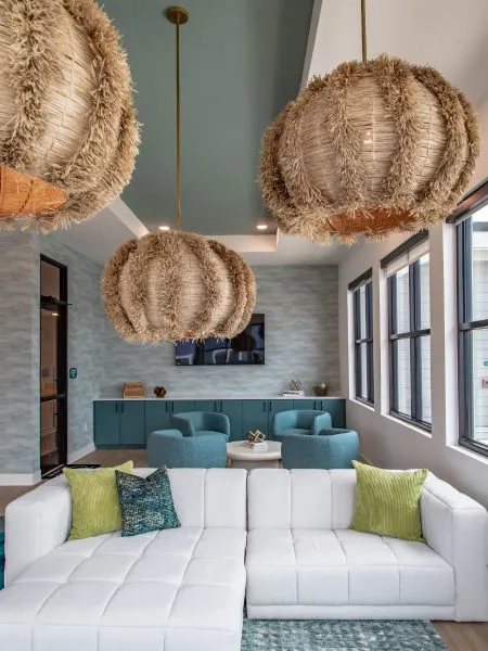 Clubroom lit by three fringed raffia globe pendants, a white tufted sectional, teal swivel chairs and a sage green ceiling