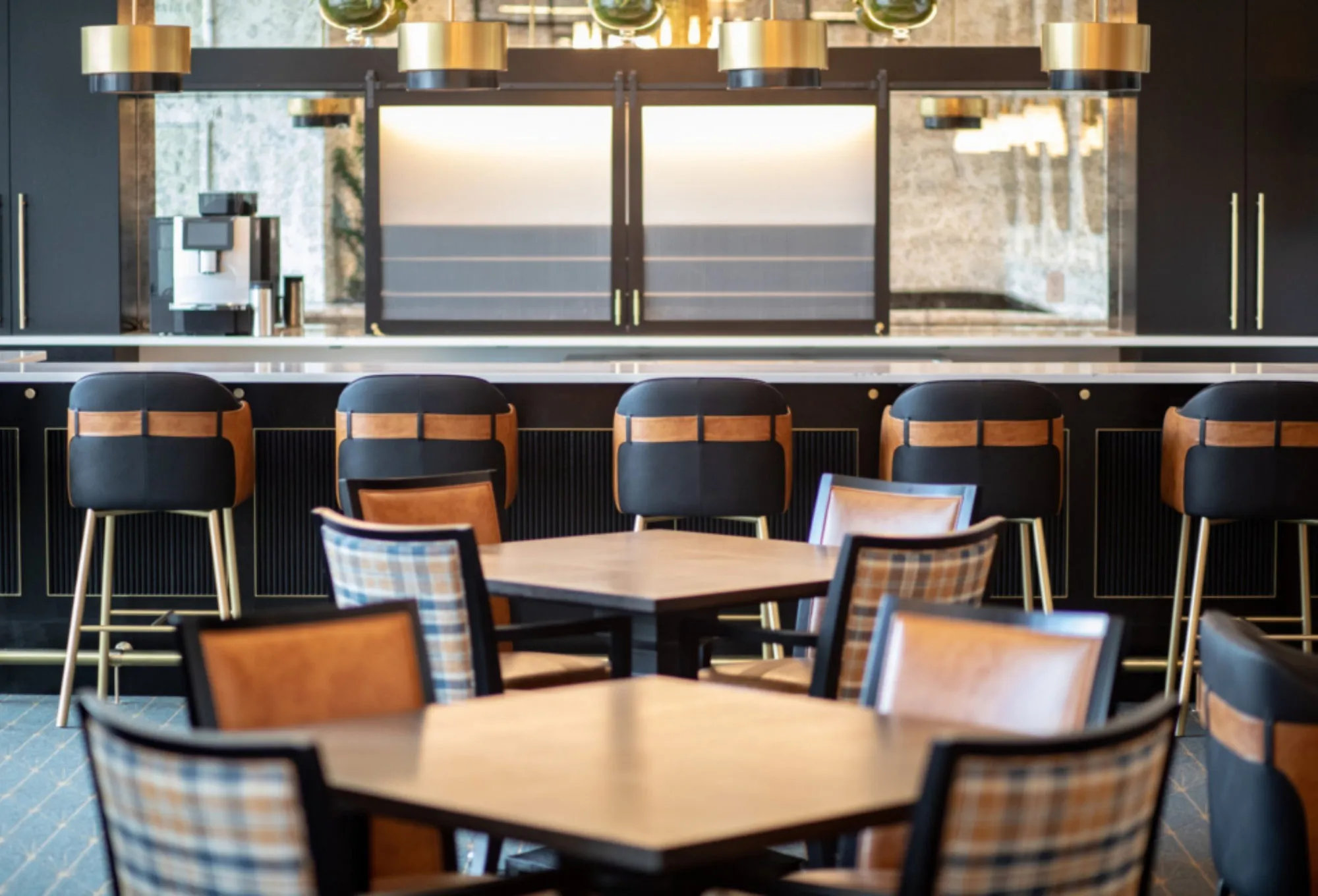 Club bar with black millwork and a mirrored back bar, brass and glass pendants and leather counter stools at wood tables
