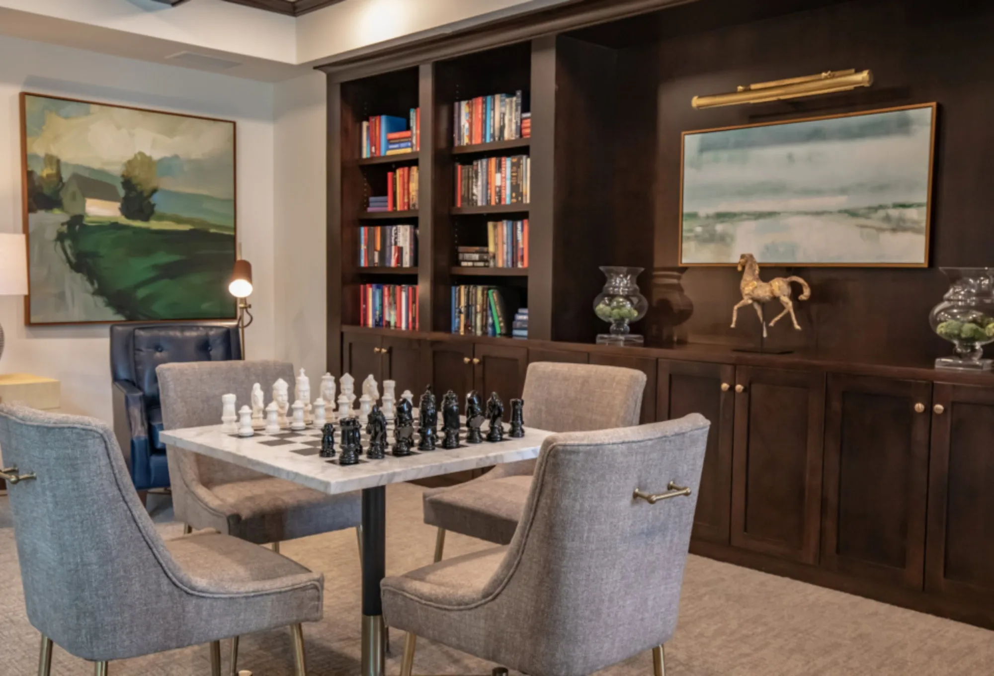 Club library with dark wood bookcases, a marble chess table set for play, grey armchairs and a brass horse sculpture