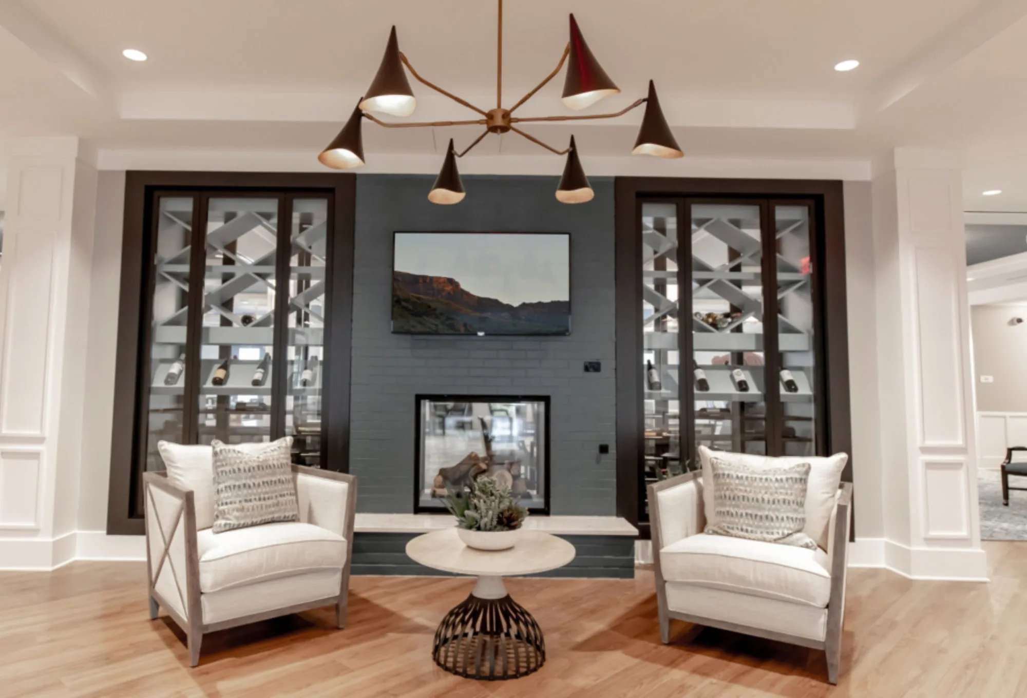 Two glass-fronted wine walls flanking a black brick fireplace, a black and brass cone chandelier above two white armchairs