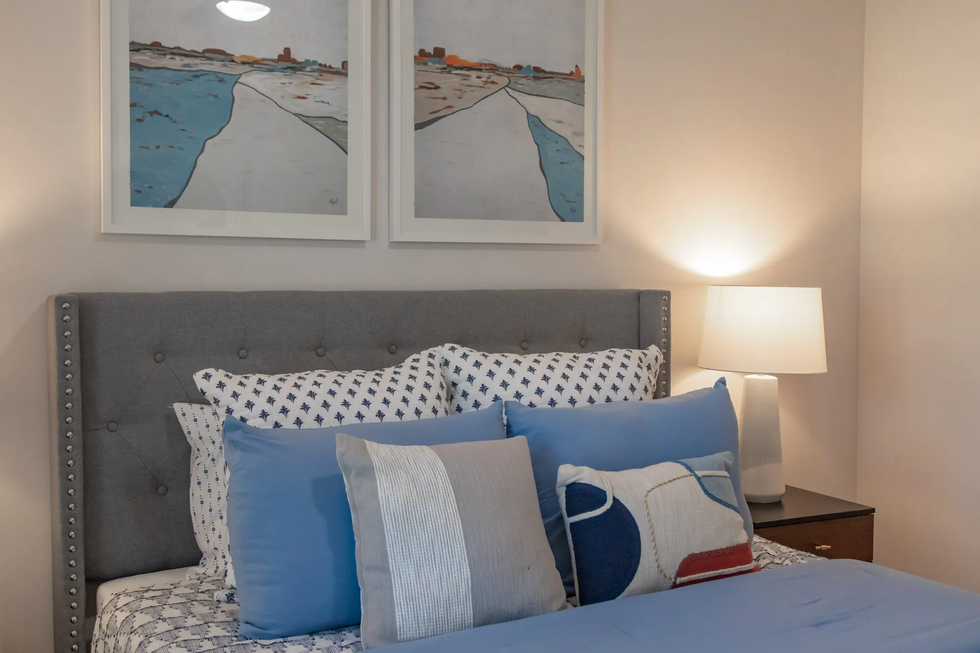 Multifamily apartment model bedroom with a grey tufted headboard, blue and patterned pillows and two framed coastal prints
