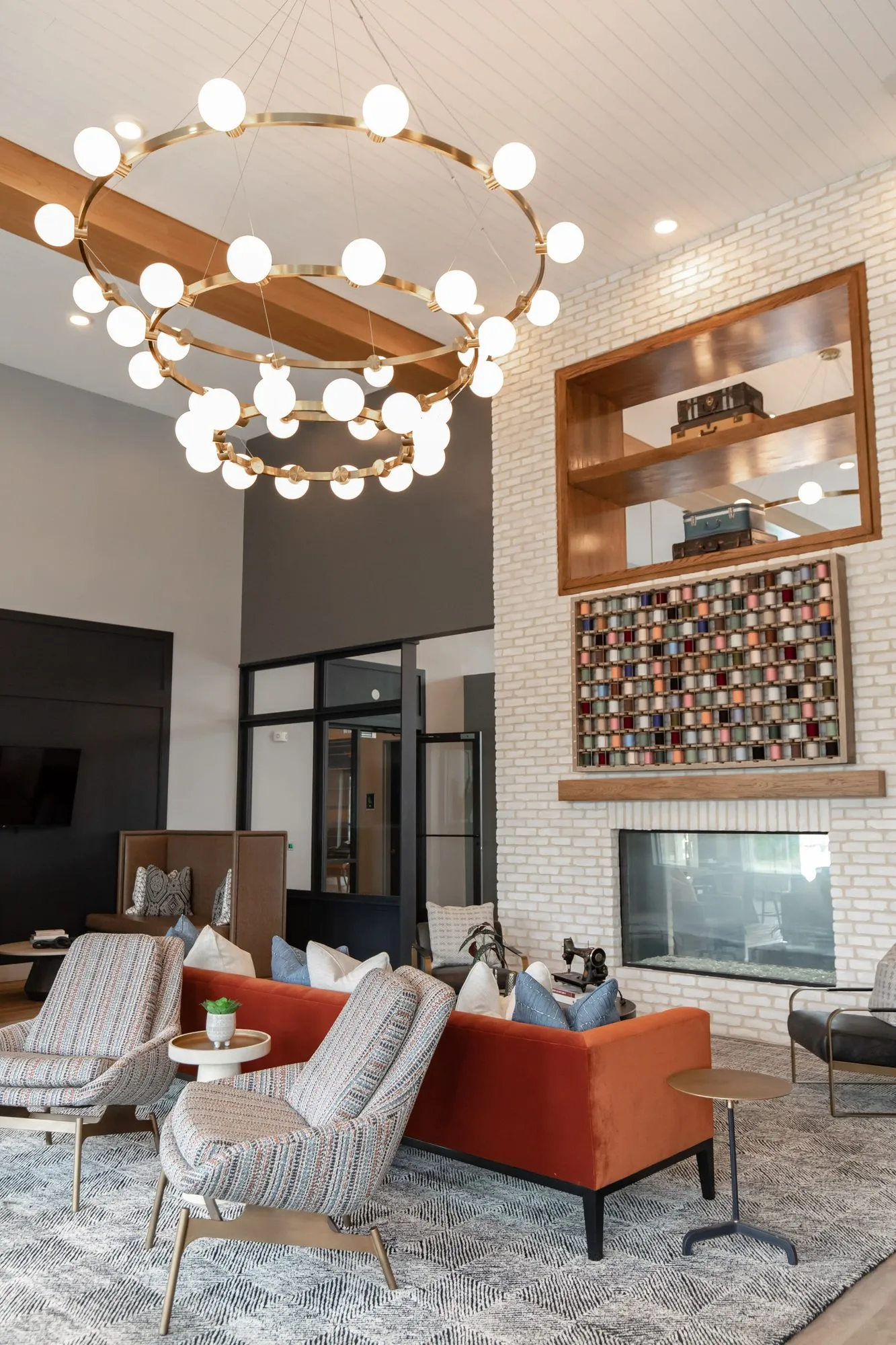 Multifamily amenity lounge under a brass double-ring globe chandelier toward a rust sofa and the white brick fireplace wall