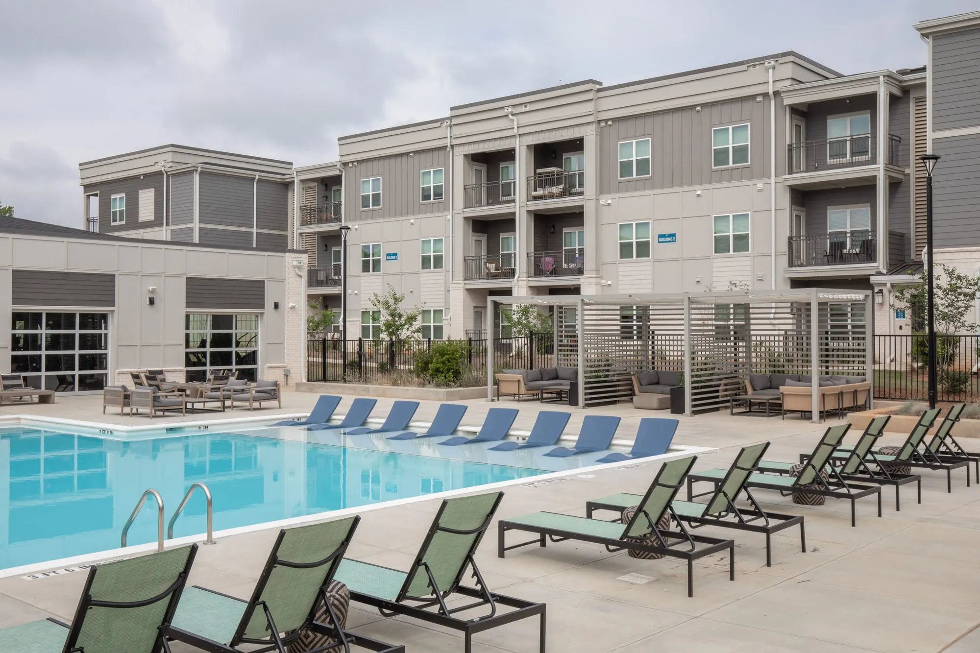 Multifamily community pool deck with green and navy chaise loungers along the water, pergola seating and apartments beyond