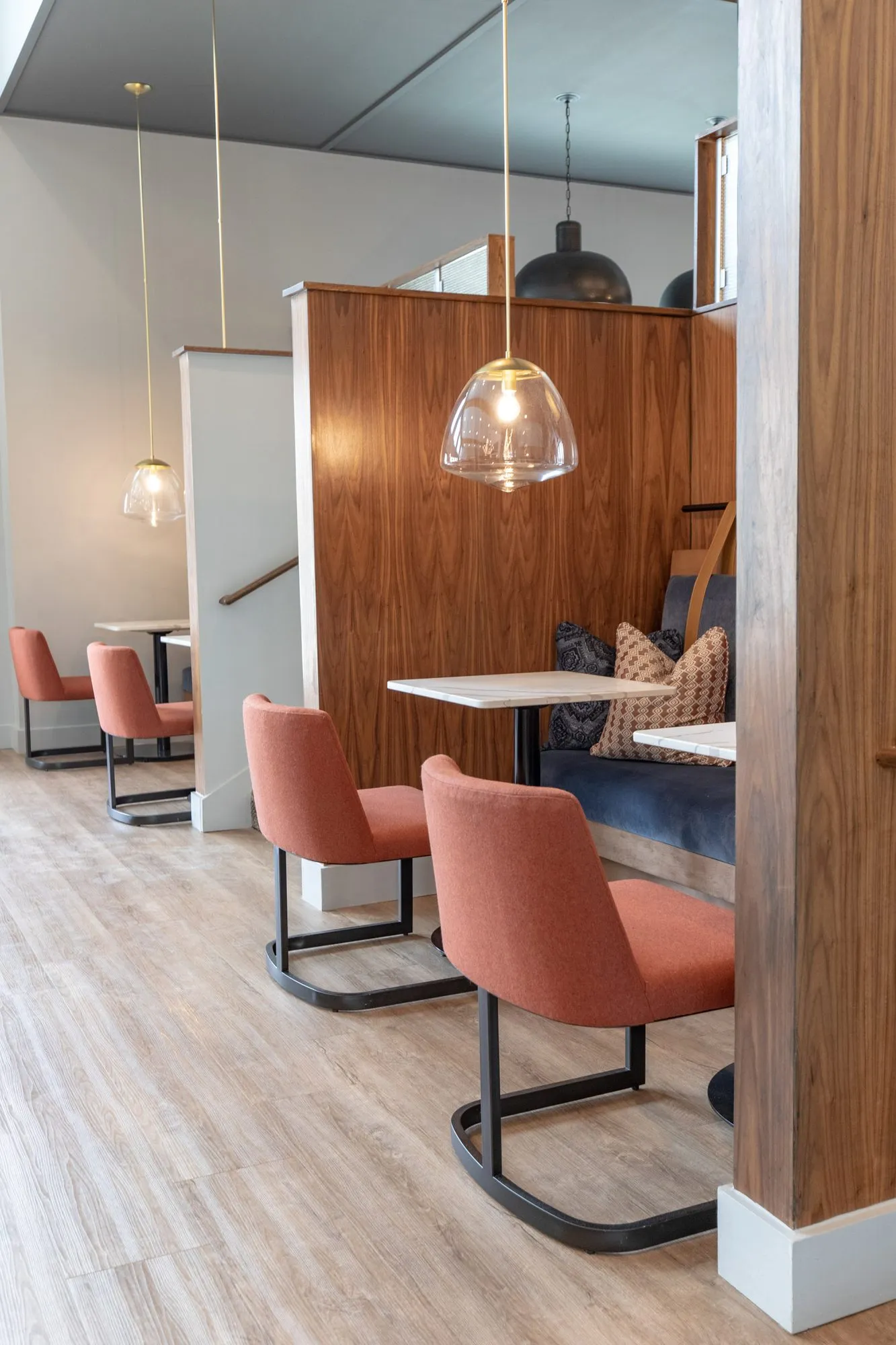 Multifamily amenity booths seen from the side, walnut partitions and rust chairs at small white tables under glass pendants