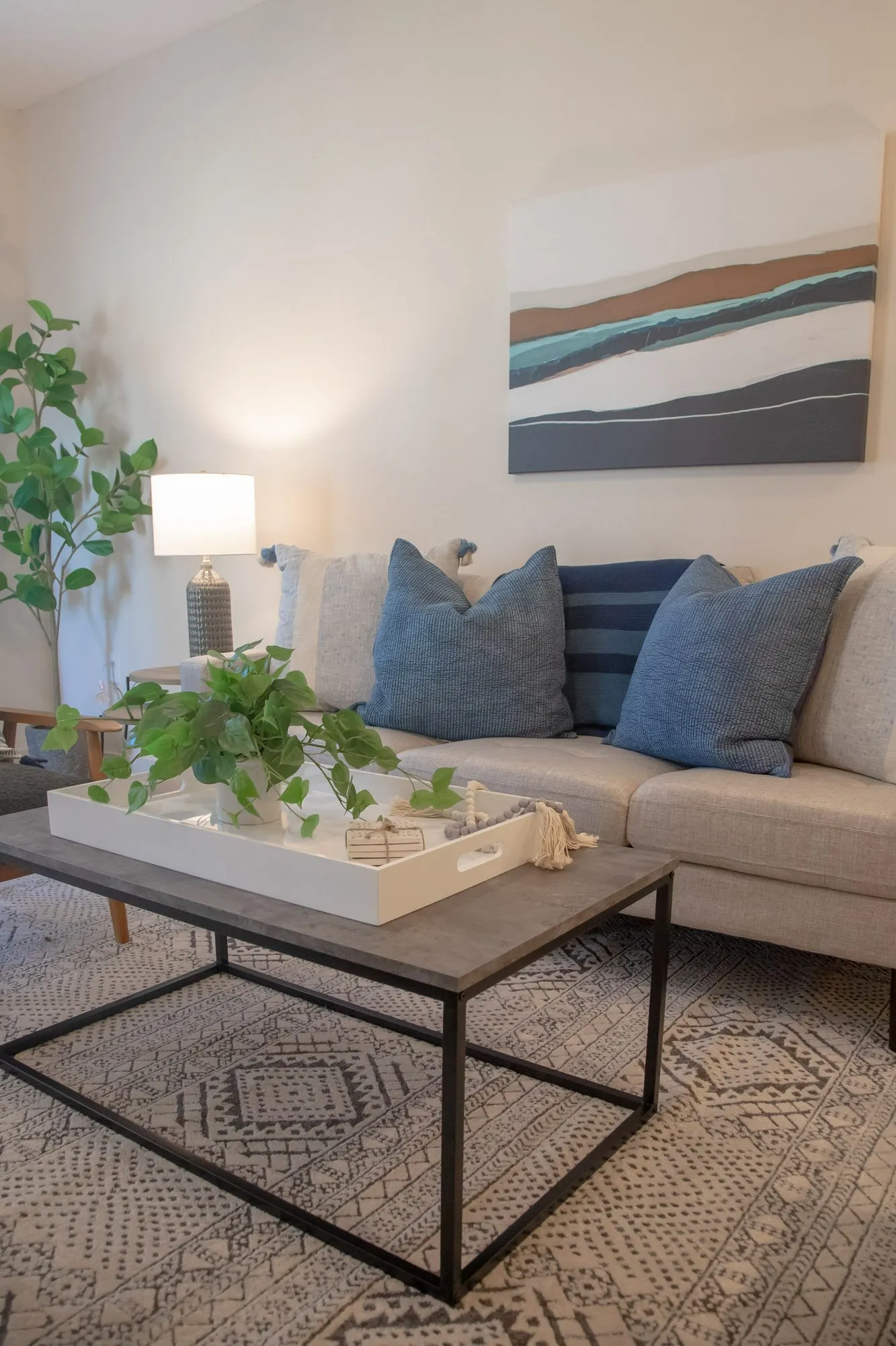 Multifamily apartment model living room with a pale sofa, blue pillows and a wood coffee table below an abstract painting