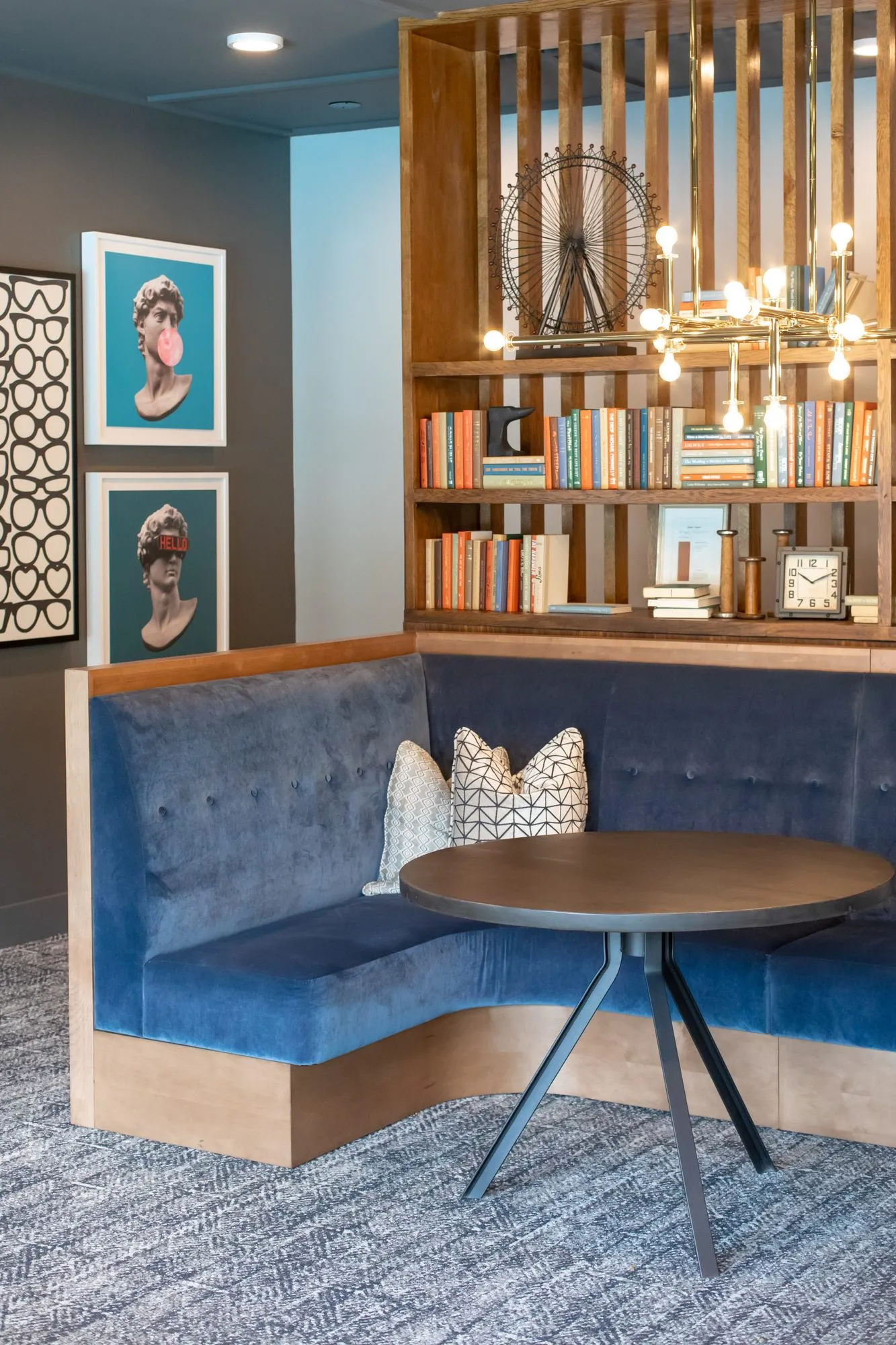 Multifamily amenity booth nook with a navy velvet corner banquette below a wood shelf screen and framed pop art busts