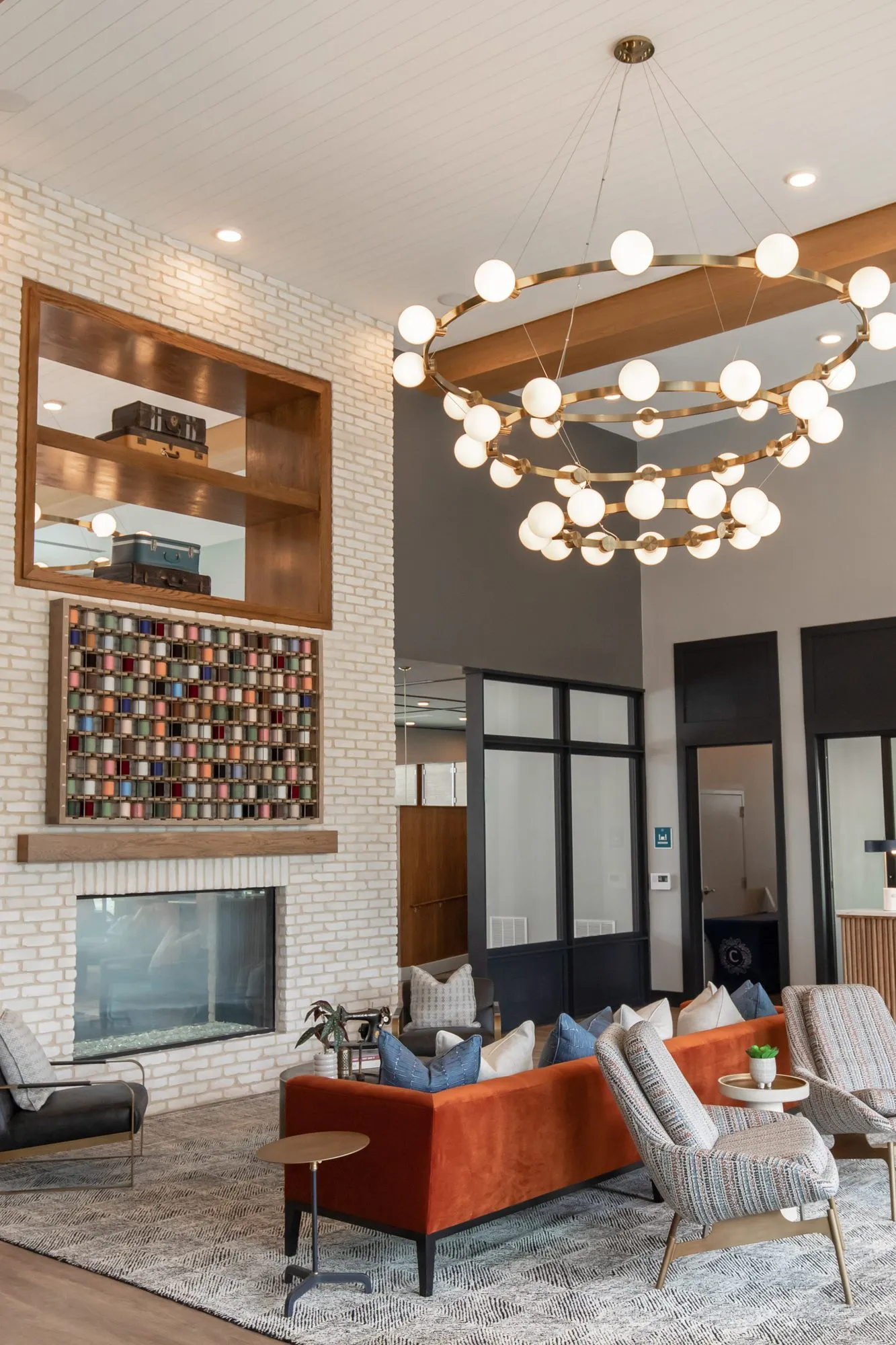 Multifamily amenity lounge with the white brick fireplace wall, spool display and wood niche, the ring chandelier above